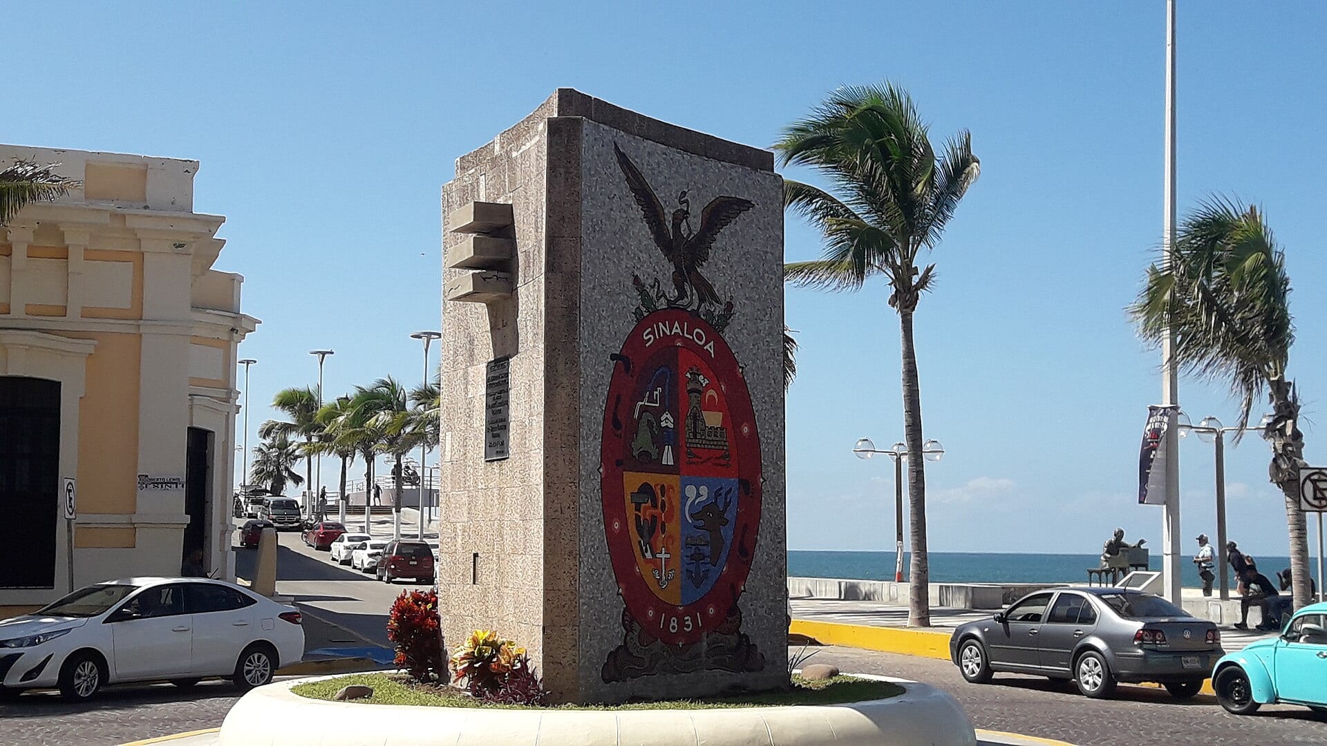 Monumento con el escudo de Sinaloa en una glorieta del malecón de Mazatlán, con palmeras y el mar al fondo.