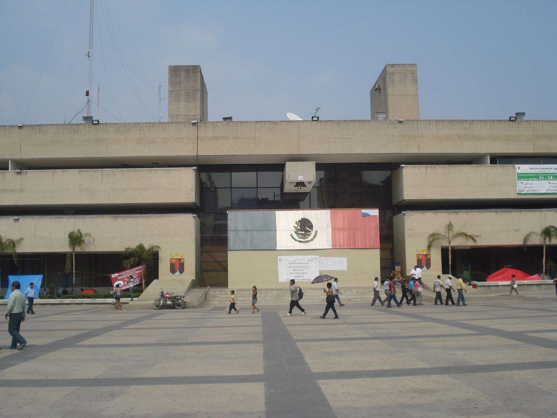 Fachada de concreto del Palacio de Gobierno de Chiapas, en Tuxtla Gutiérrez, con una bandera pintada en el acceso y personas cruzando la plaza.