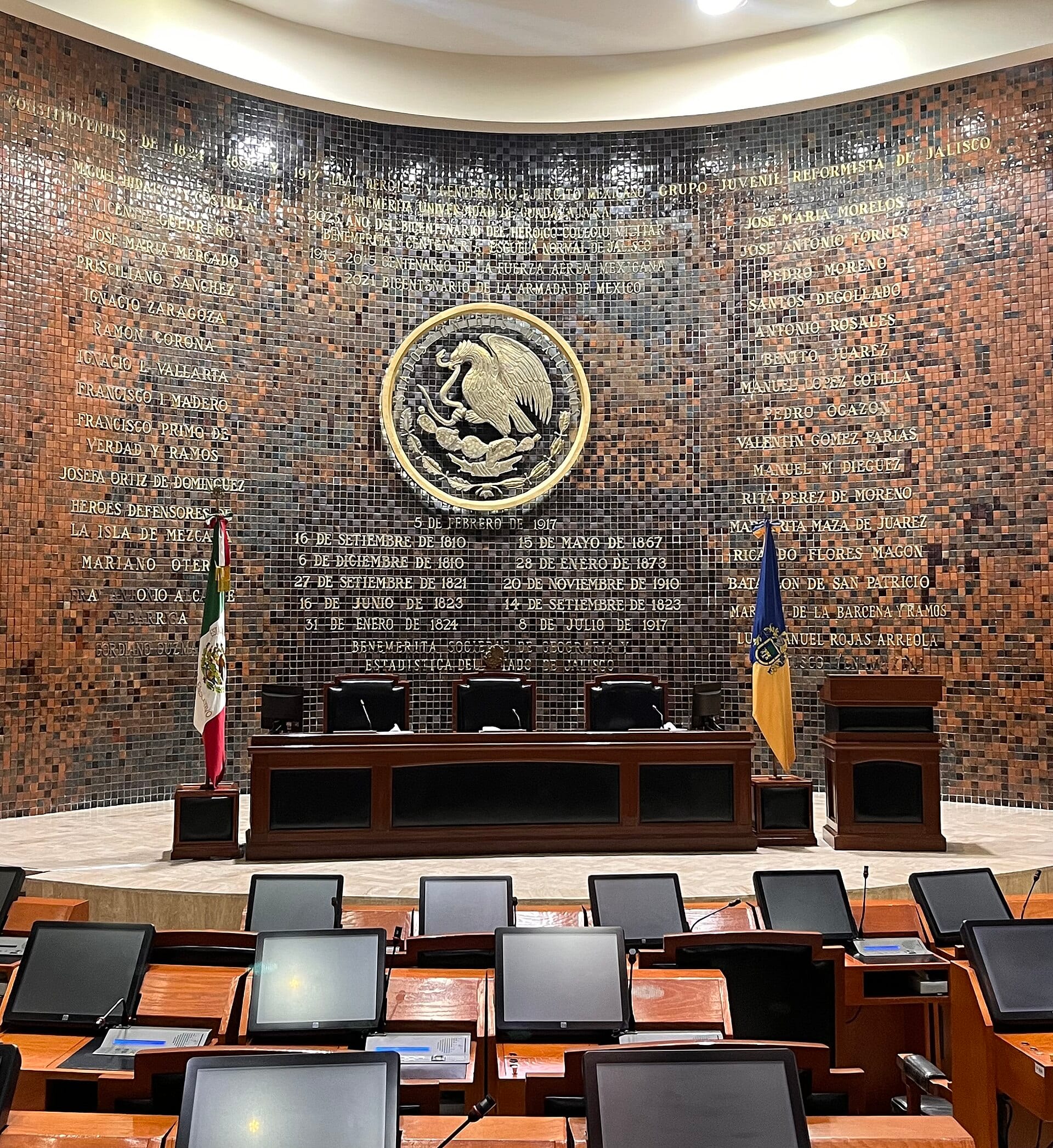 Sala de sesiones del Congreso de Jalisco, con el escudo nacional sobre un muro de mosaico, la mesa directiva entre dos banderas y las curules en primer plano.