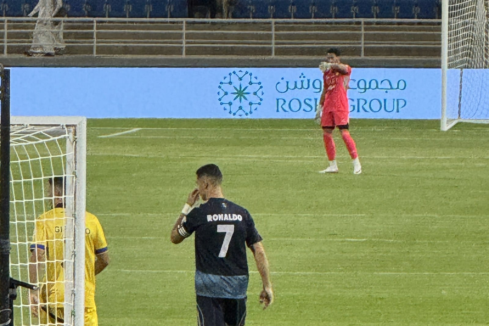 Cristiano Ronaldo, seen from behind in Al Nassr's number 7 shirt, raises a hand to his face on a grass pitch; a goalkeeper in red stands in the background.