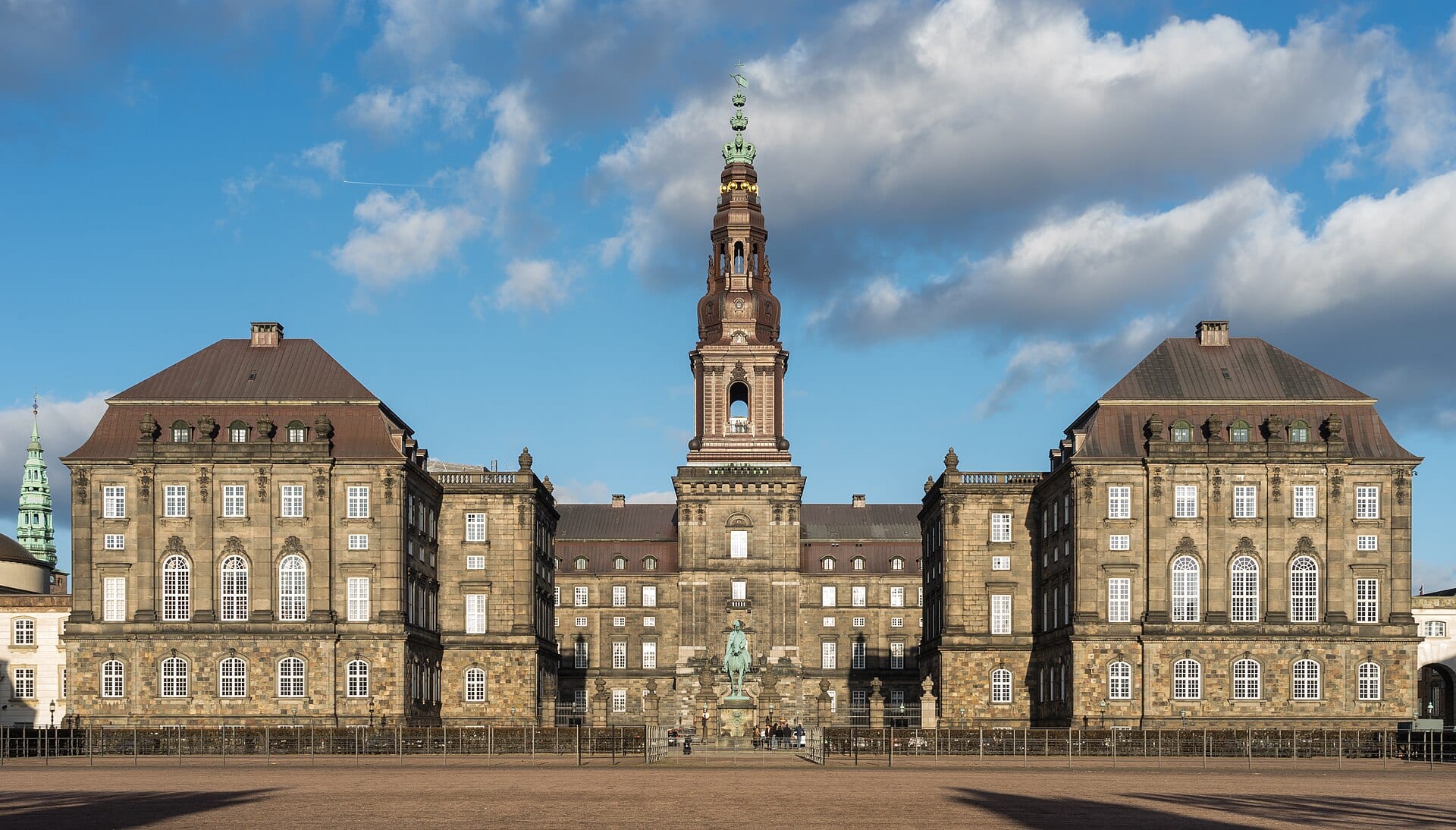 The stone facade of Christiansborg Palace in Copenhagen, with a central tower under a blue sky with clouds.