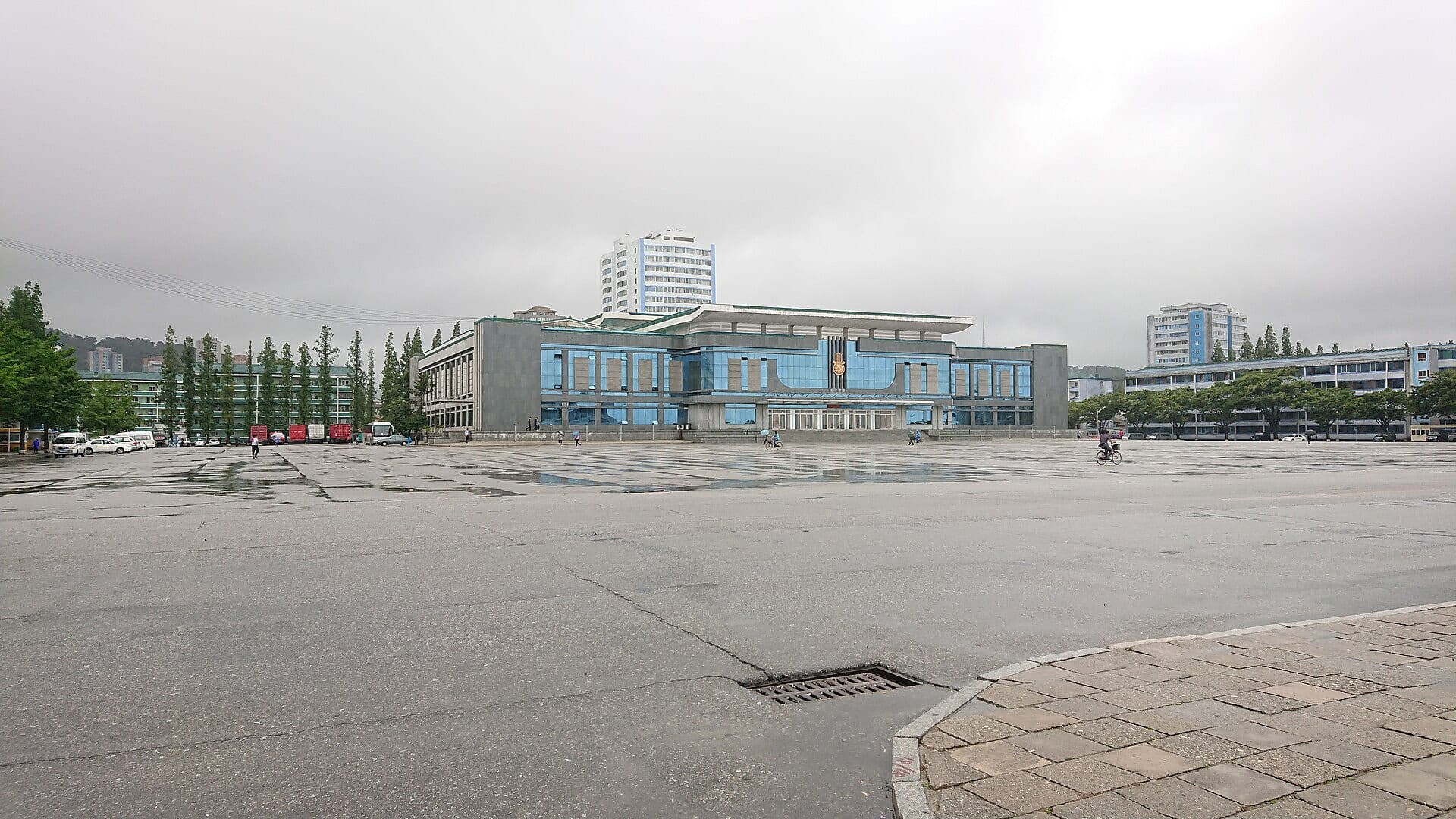 A wide, wet plaza in Wonsan with a blue-fronted building behind it and a cyclist crossing under a cloudy sky.