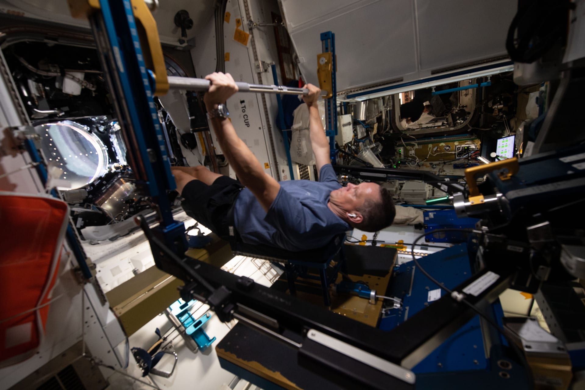 Un astronauta acostado levanta una barra en un dispositivo de ejercicio de resistencia dentro de la Estación Espacial Internacional.
