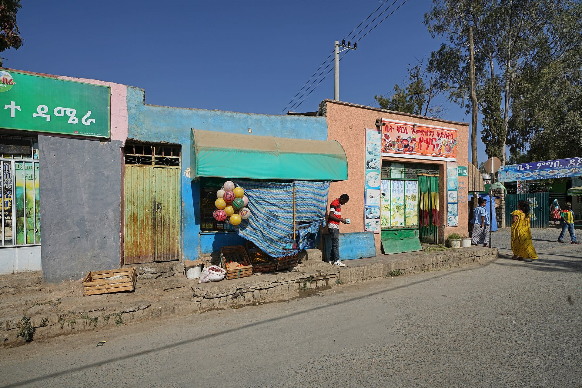 A street in Mekelle with blue and salmon-colored storefronts, a fruit stand under a green awning and people walking by.