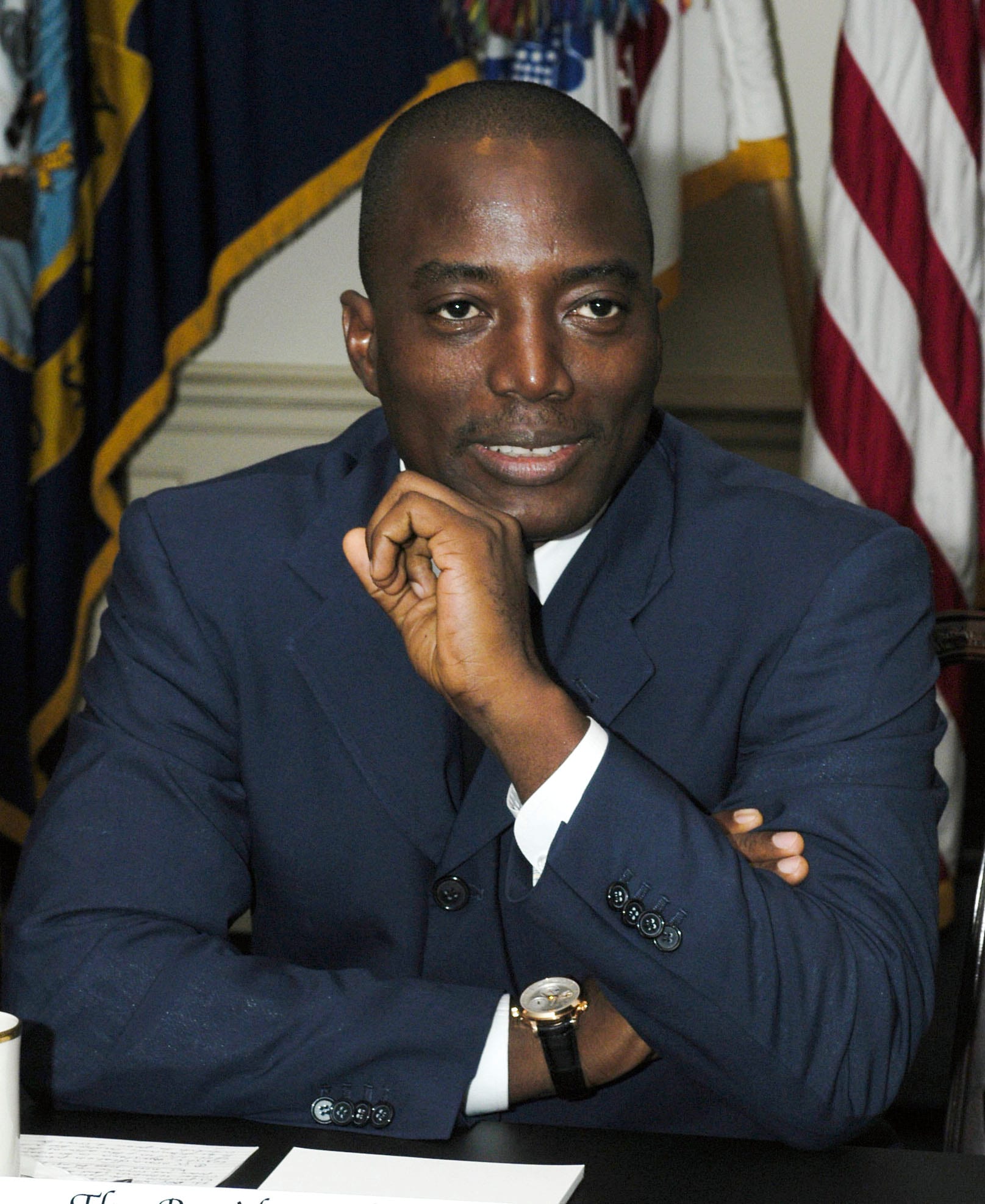 Portrait of Joseph Kabila in a blue suit, seated at a table with his chin resting on his hand and flags behind him.