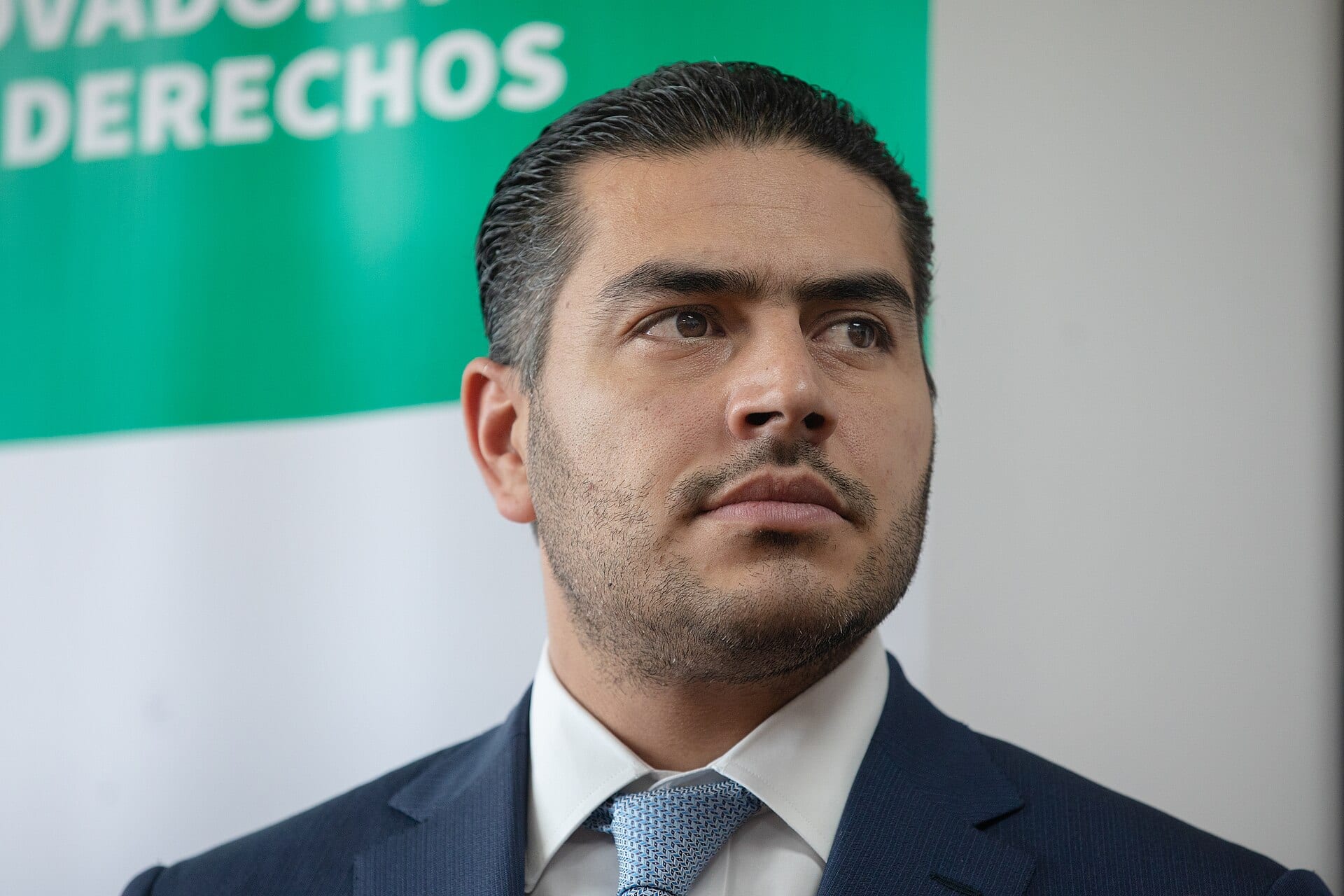 Primer plano de Omar García Harfuch, de saco azul y corbata, con la mirada hacia un lado frente a un fondo verde y blanco.