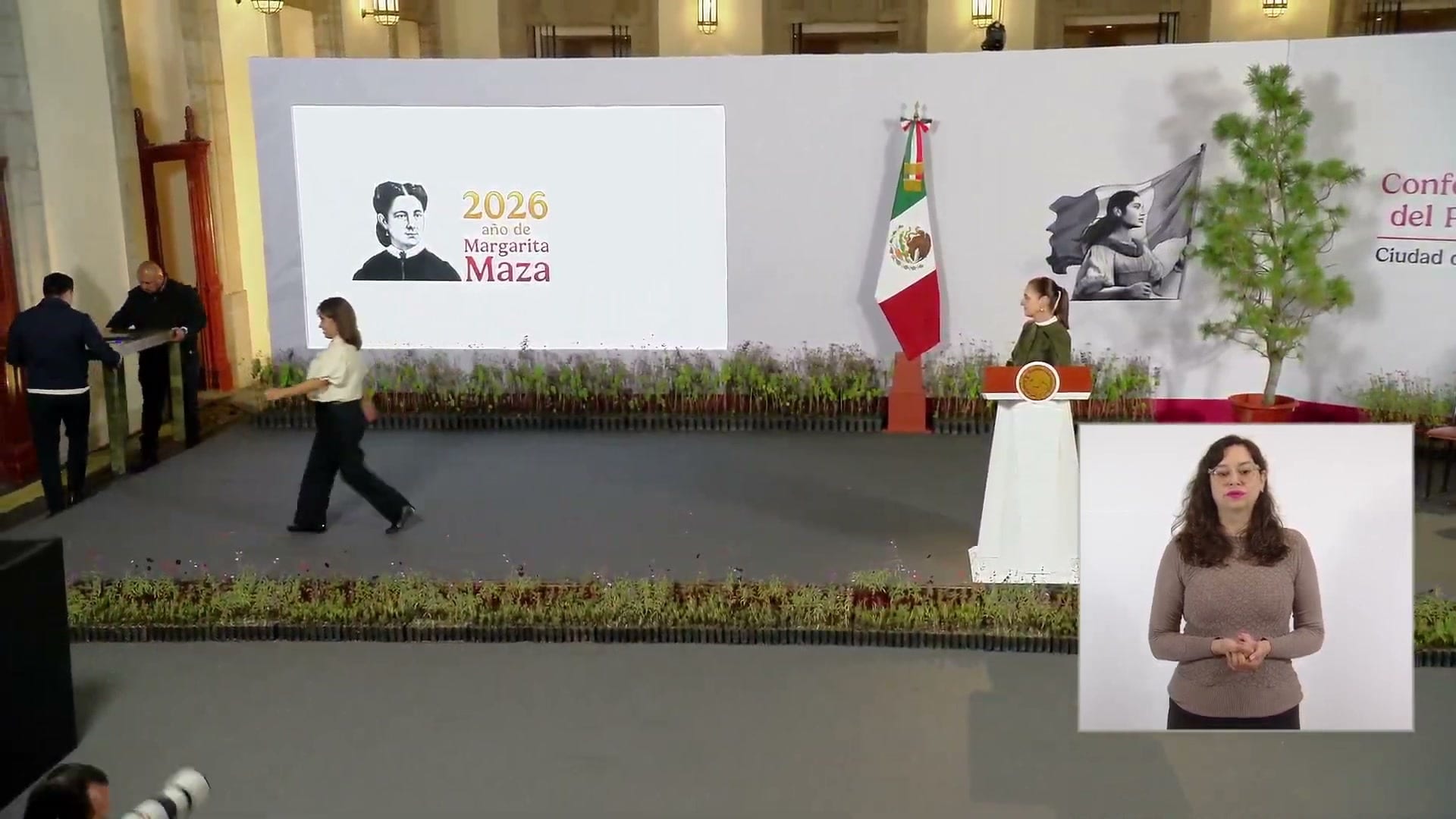 Escenario de la conferencia matutina con el podio, una lámina del año de Margarita Maza, hileras de plantas y un árbol en maceta.