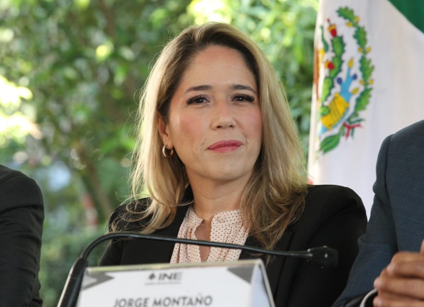 La consejera electoral Frida Gómez, de cabello rubio y saco negro, sentada ante un micrófono junto a una bandera de México.