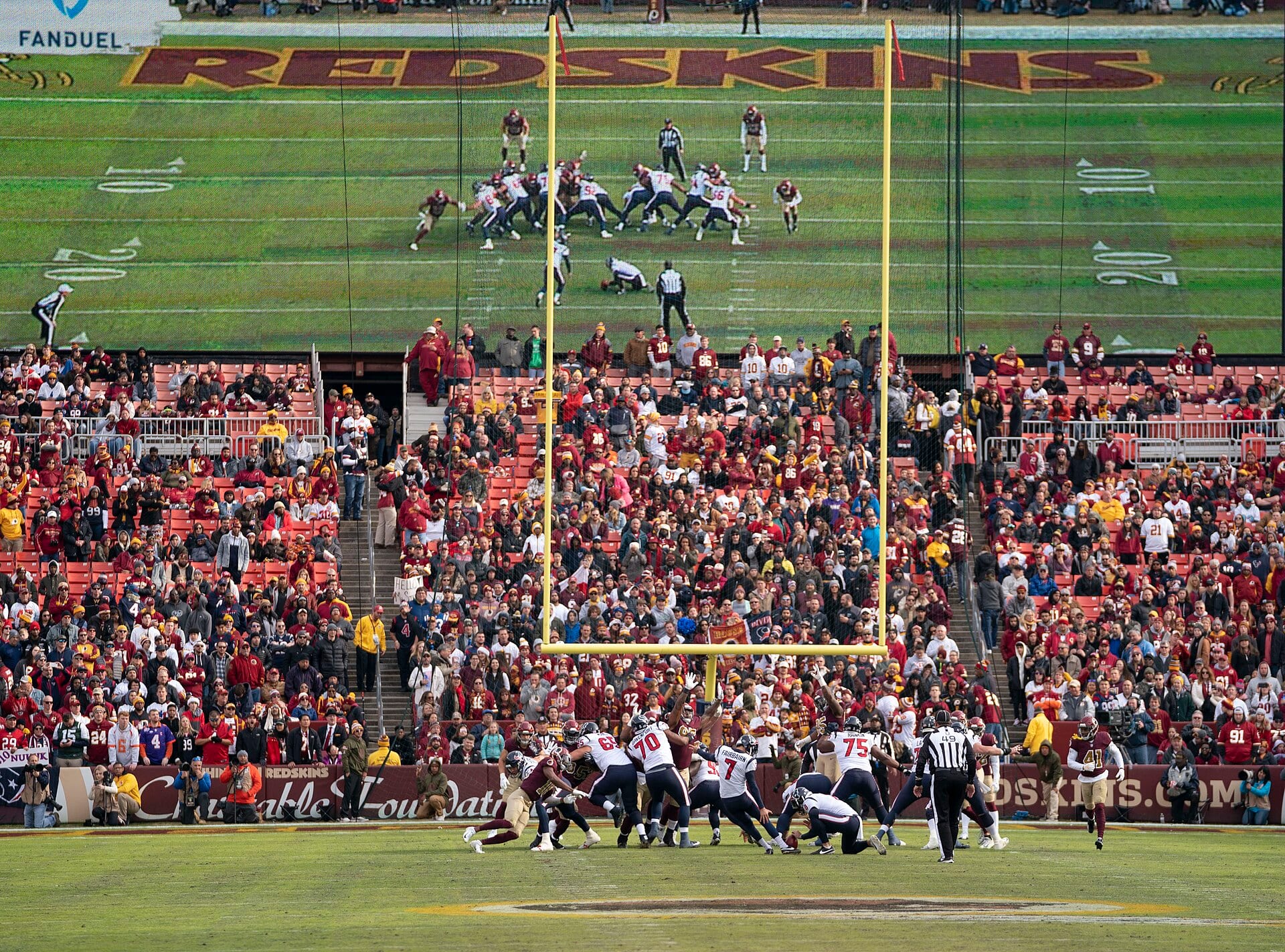 El pateador número 7 de los Texans golpea el balón entre la línea ofensiva, con las tribunas llenas y una pantalla gigante al fondo del estadio.