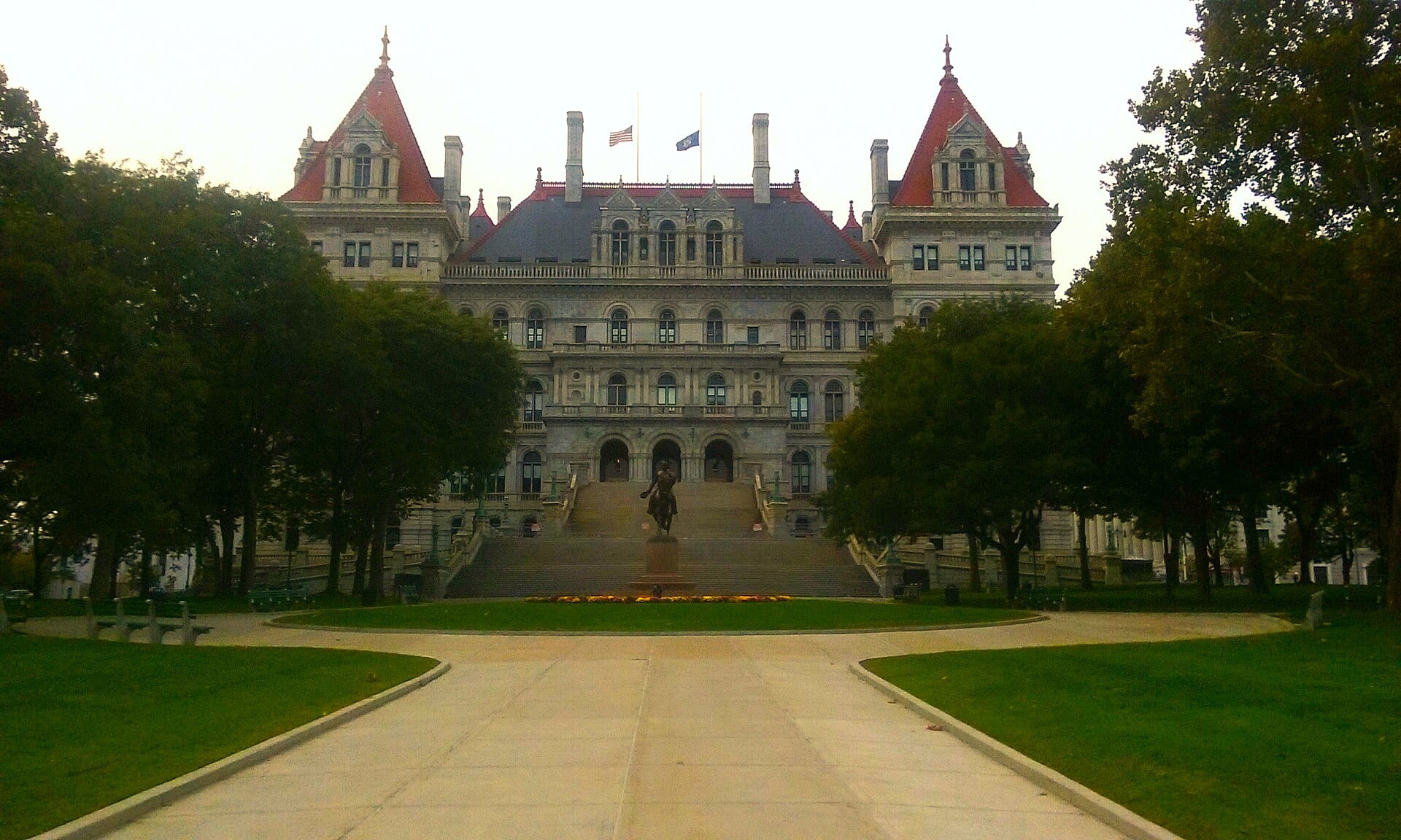 The New York State Capitol, a stone building with red-roofed towers, seen from a path between trees and lawn.