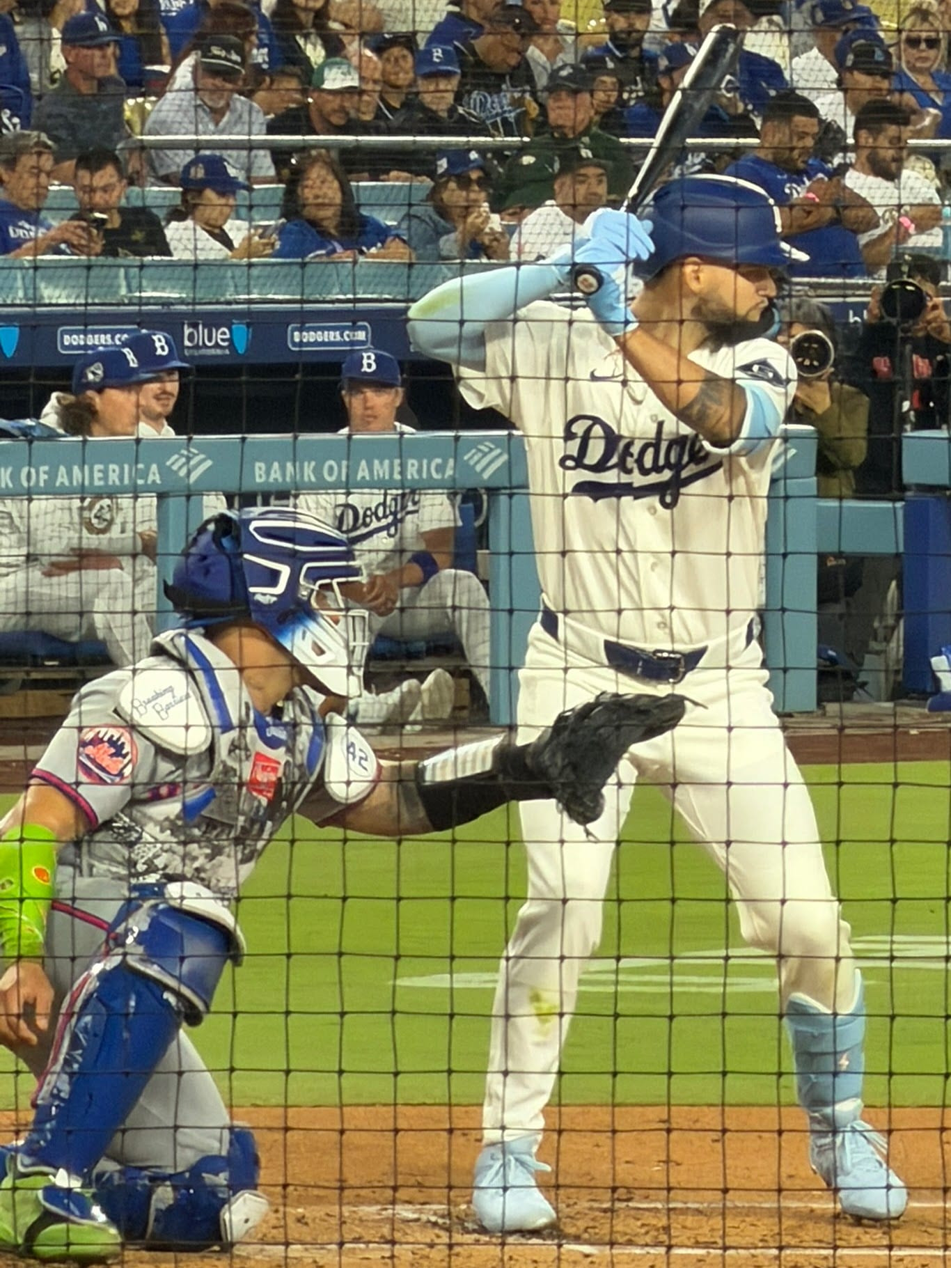 A Dodgers batter in a white uniform holds the bat high while a crouching catcher waits behind him, seen through the protective netting.