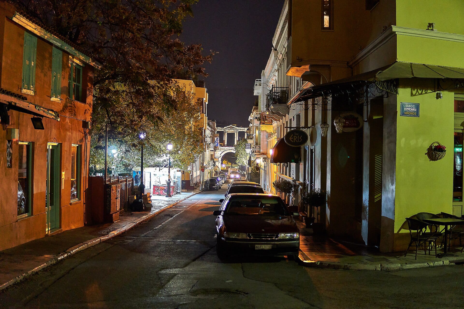 Una calle empedrada y angosta de Plaka de noche, con autos estacionados y el Arco de Adriano iluminado al fondo.