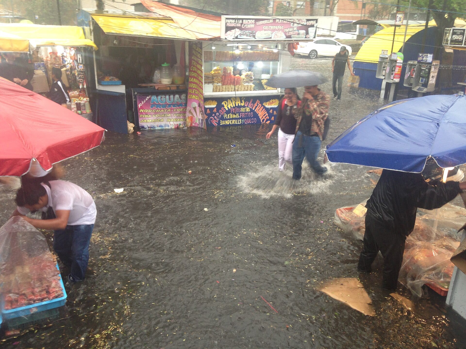Vendedores y transeúntes cruzan una calle inundada junto a puestos de comida, algunos con paraguas, durante un aguacero en la Ciudad de México.