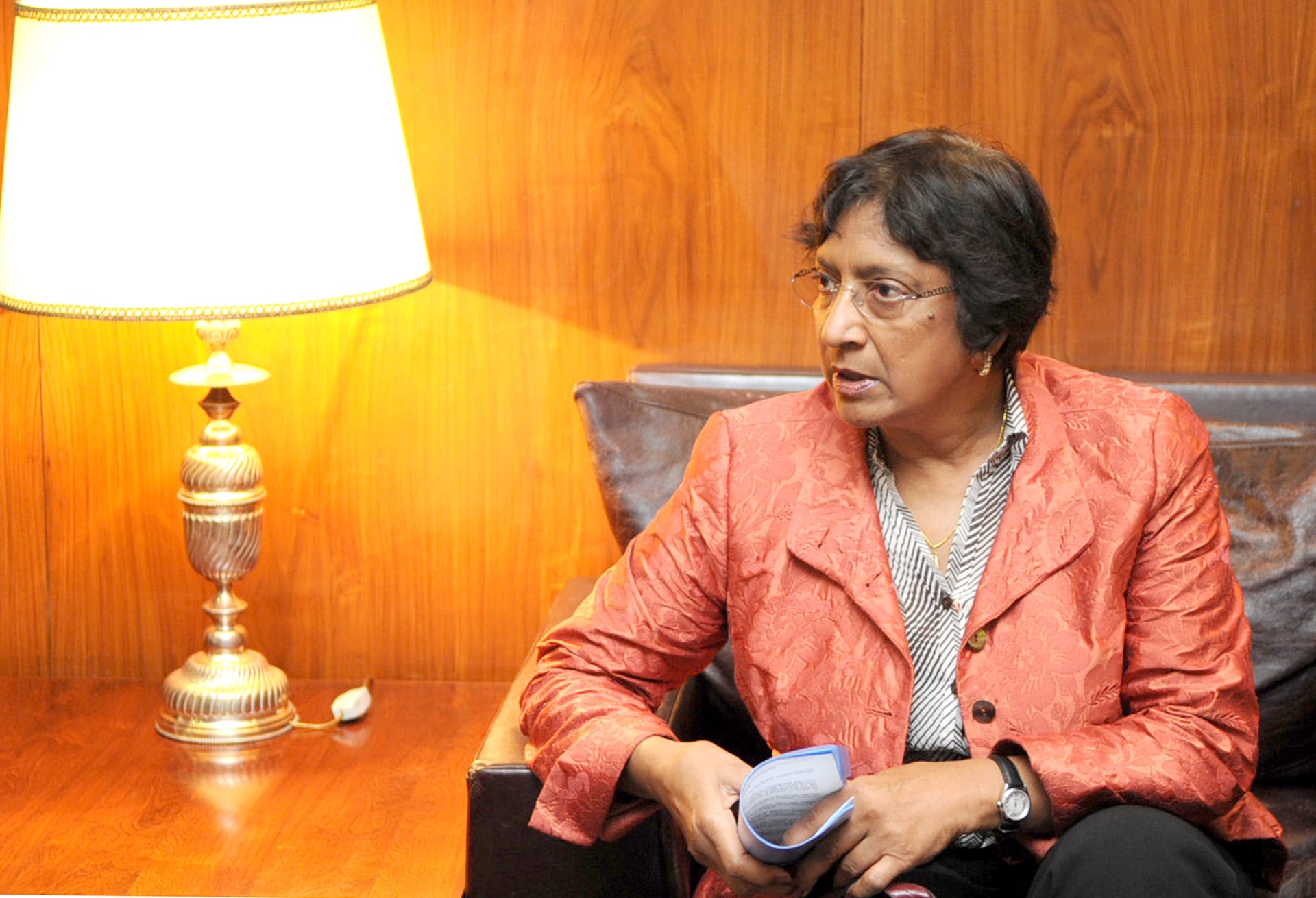 A woman with short dark hair, glasses and a rust-colored jacket looks to the side from an armchair next to a lamp.
