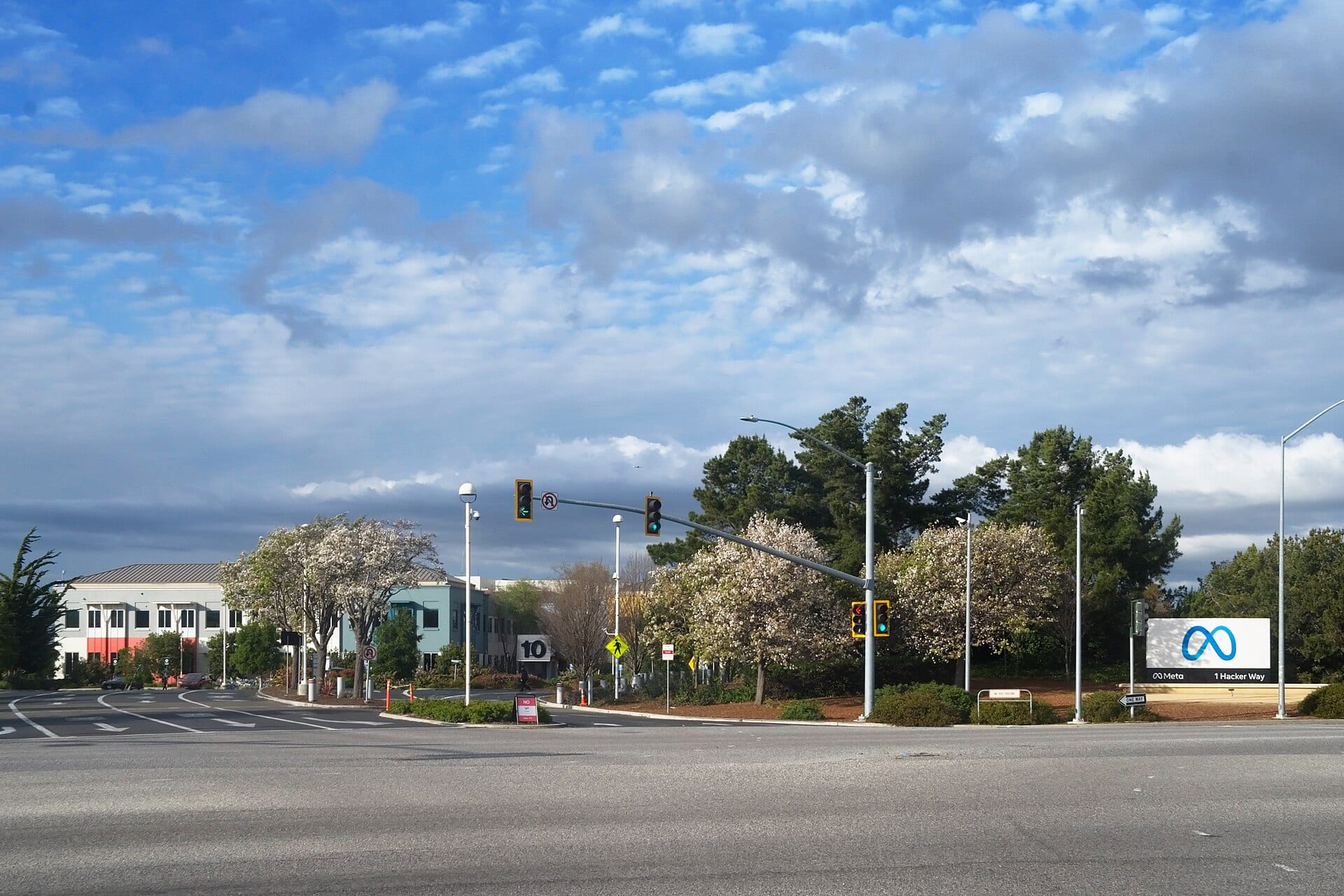 Entrada del complejo de Meta en Menlo Park, California: un cruce con semáforos, árboles en flor y el letrero de Meta con la dirección 1 Hacker Way.