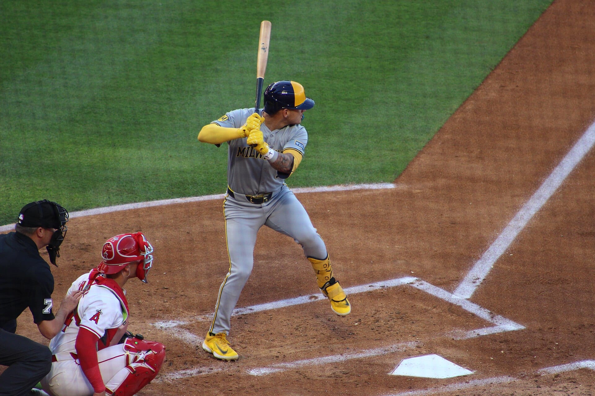 Un bateador de los Brewers con casco azul y amarillo y uniforme gris espera el lanzamiento junto al plato, con el árbitro y el receptor atrás.