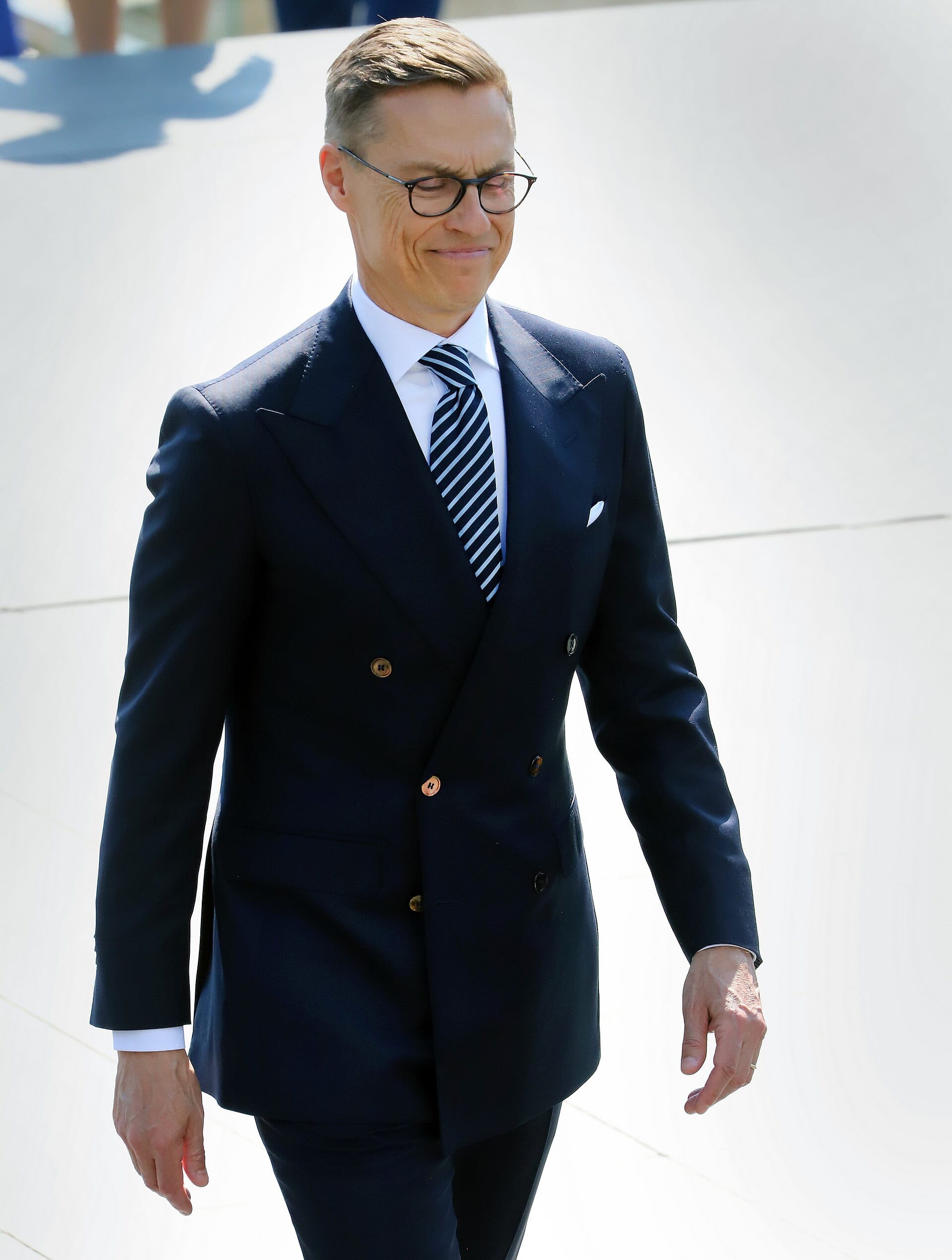 Alexander Stubb, in a navy suit and striped tie, walks with a restrained smile across a white surface.