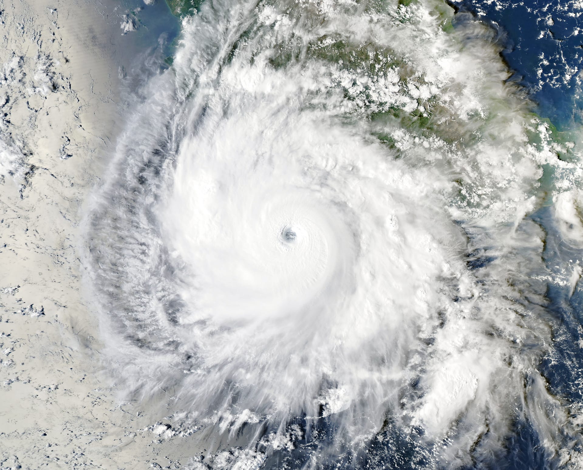 Vista satelital de un huracán con un ojo pequeño al centro y bandas de nubes blancas en espiral sobre el océano, con la costa al fondo.