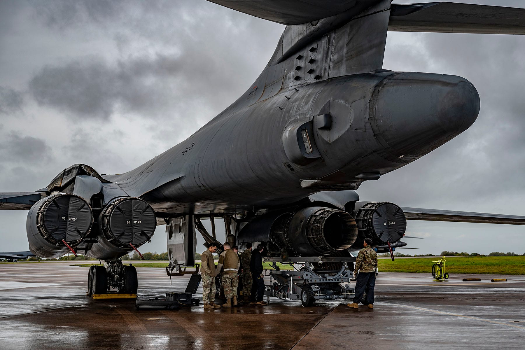 Un grupo de militares de uniforme camuflado trabaja bajo la cola de un bombardero B-1 Lancer gris sobre una pista mojada, con cielo nublado de fondo.
