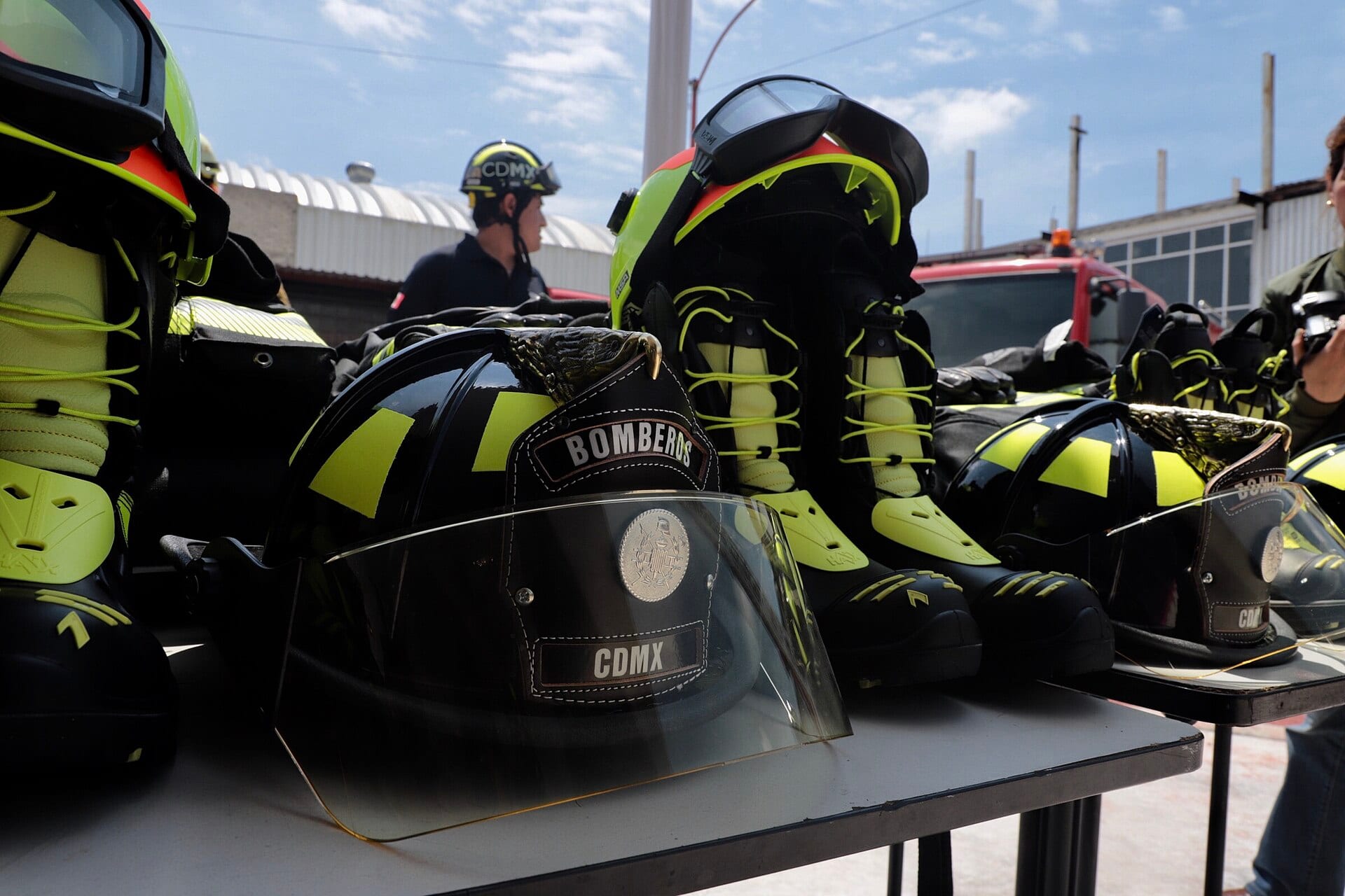 Cascos negros con la leyenda Bomberos CDMX y botas con franjas amarillas fosforescentes sobre una mesa, con un bombero al fondo.