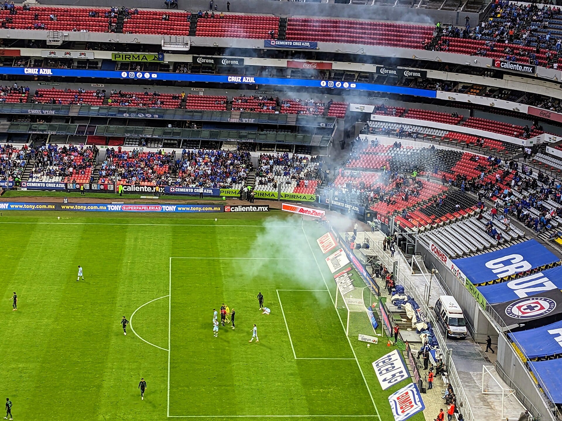 Vista elevada de un partido de Liga MX con humo sobre un costado de la cancha, jugadores dispersos y mantas azules con el escudo de Cruz Azul en las gradas.