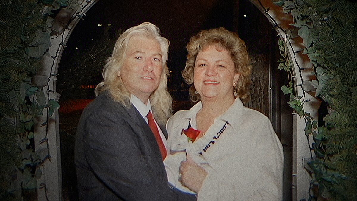 Un hombre de cabello largo y rubio, con traje gris y corbata roja, posa junto a una mujer de cabello rizado bajo un arco de follaje, de noche.