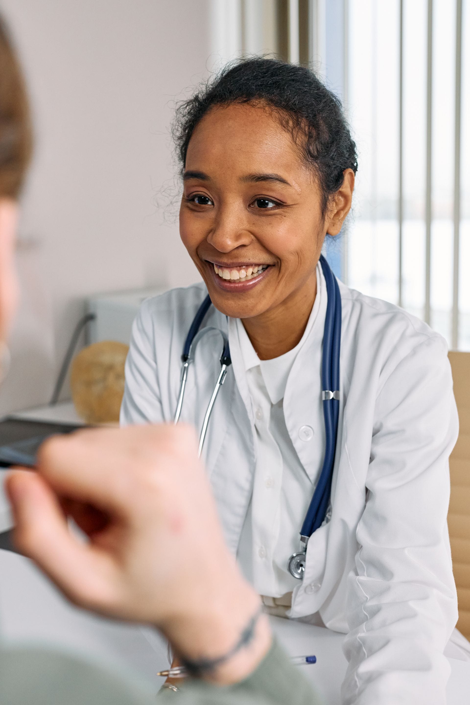 Una médica con bata blanca y estetoscopio sonríe mientras conversa con un paciente en un consultorio.