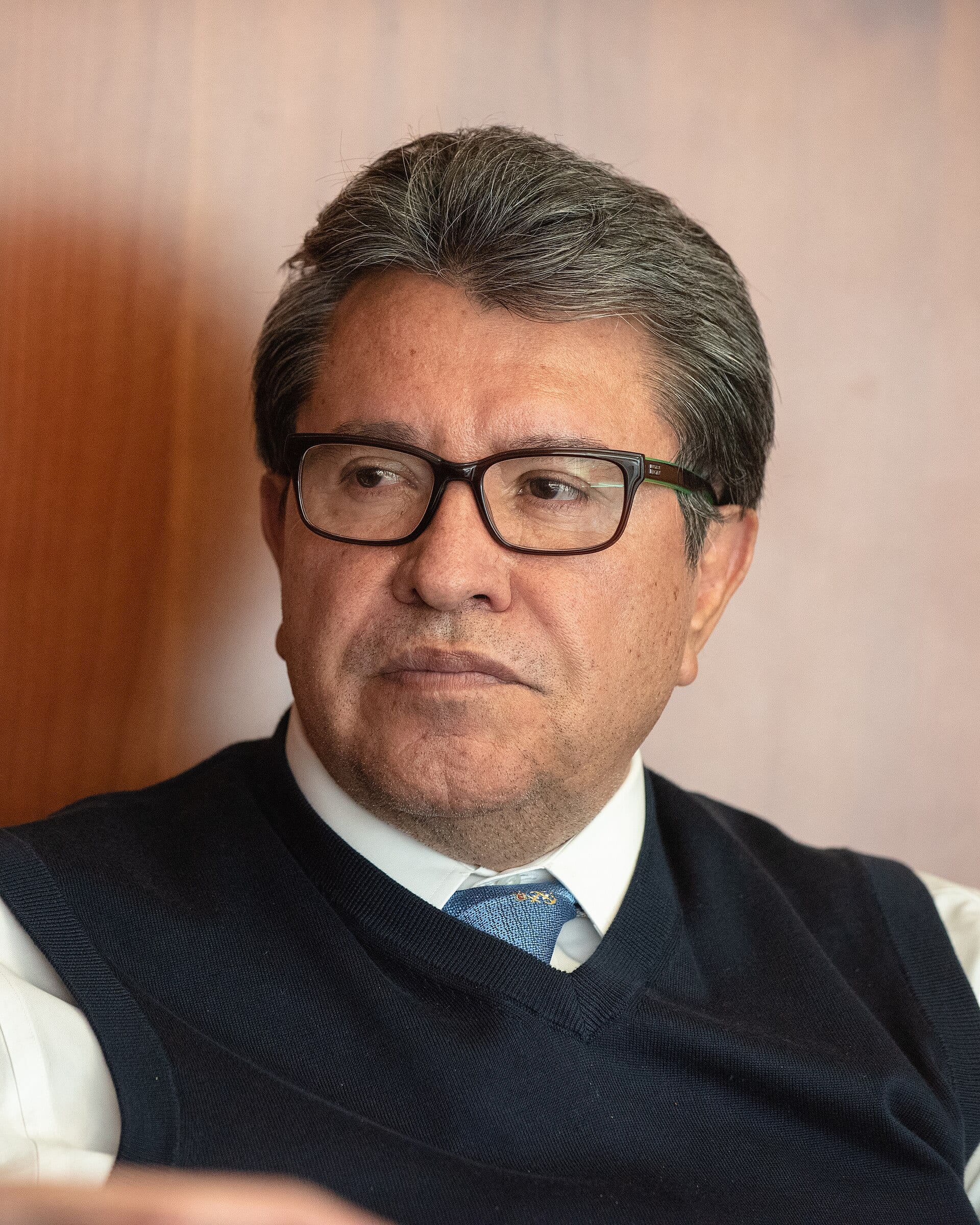 Un hombre de lentes de pasta, chaleco azul marino y corbata azul mira hacia un lado durante una entrevista.