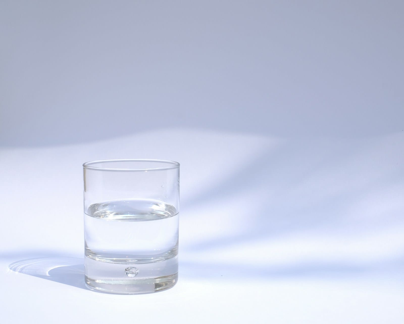 Un vaso transparente con agua hasta la mitad sobre una superficie blanca, con sombras suaves al fondo.