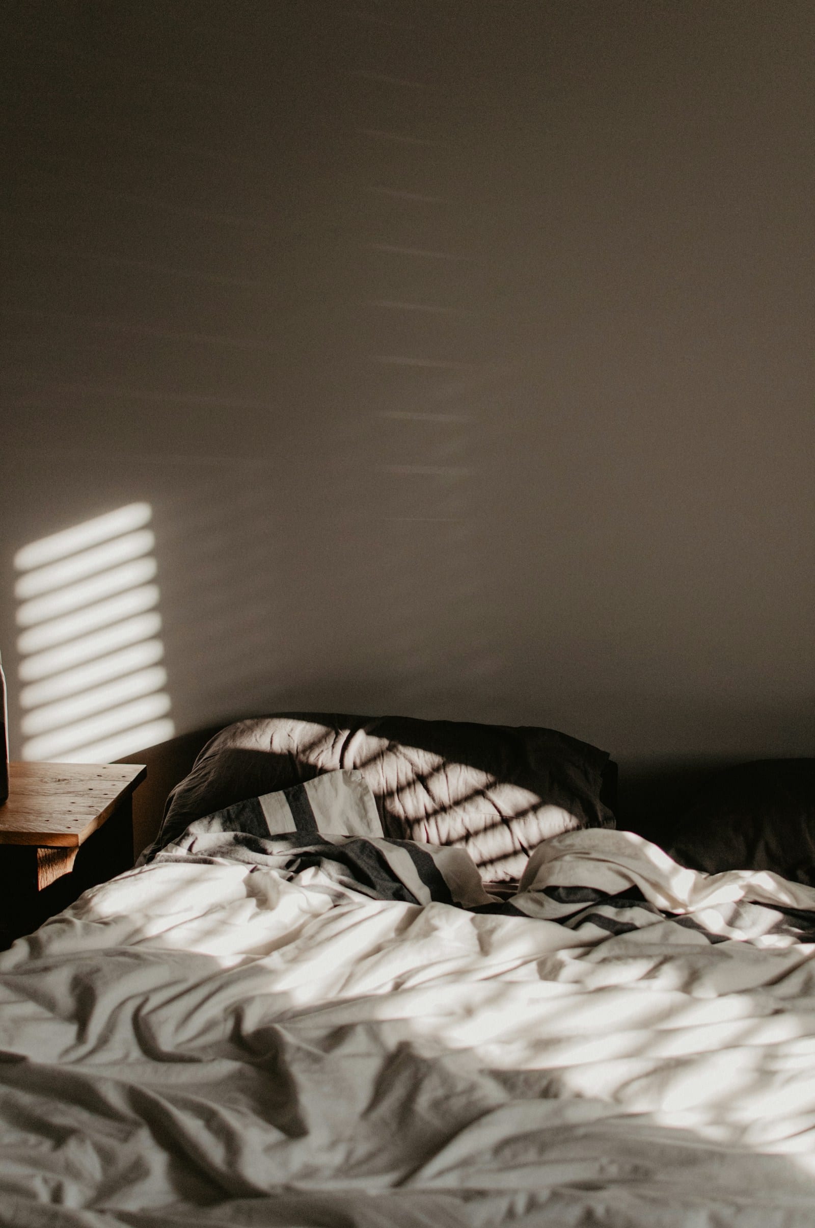 Una cama deshecha con sábanas blancas y almohadas oscuras, con franjas de luz de una persiana proyectadas sobre la pared.