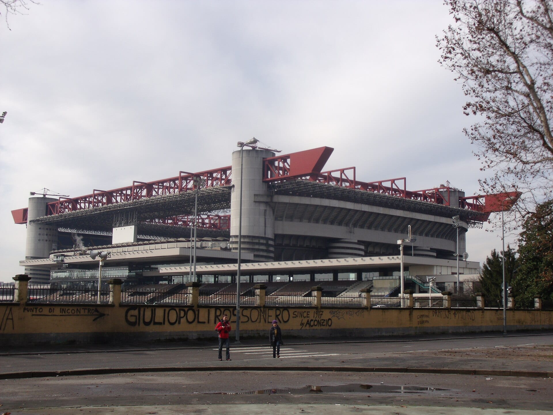 Exterior of the Giuseppe Meazza stadium, with its concrete towers and red roof beams, seen from the street.