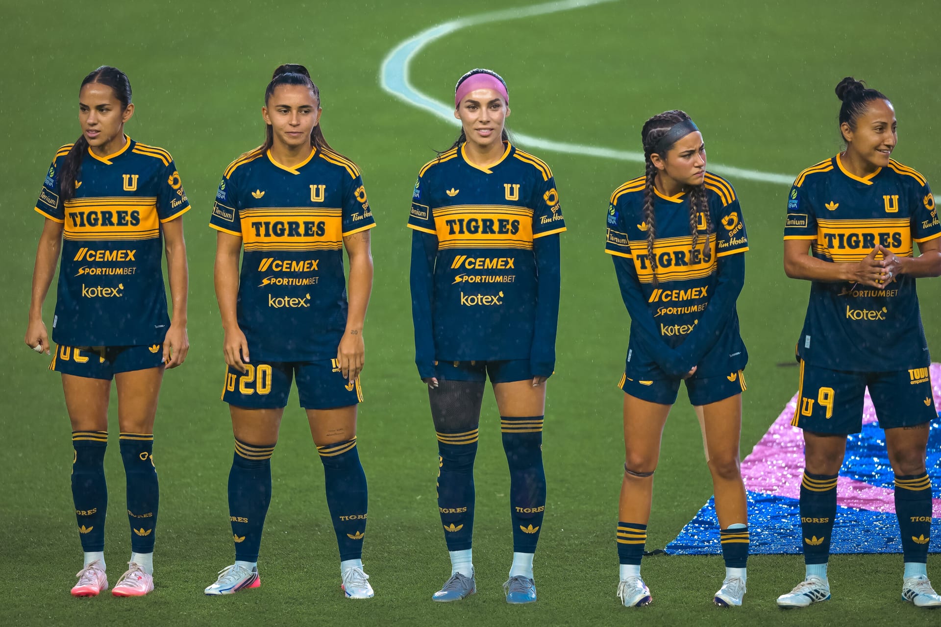 Cinco jugadoras de Tigres Femenil con el uniforme azul y dorado, alineadas sobre el pasto bajo una ligera lluvia.
