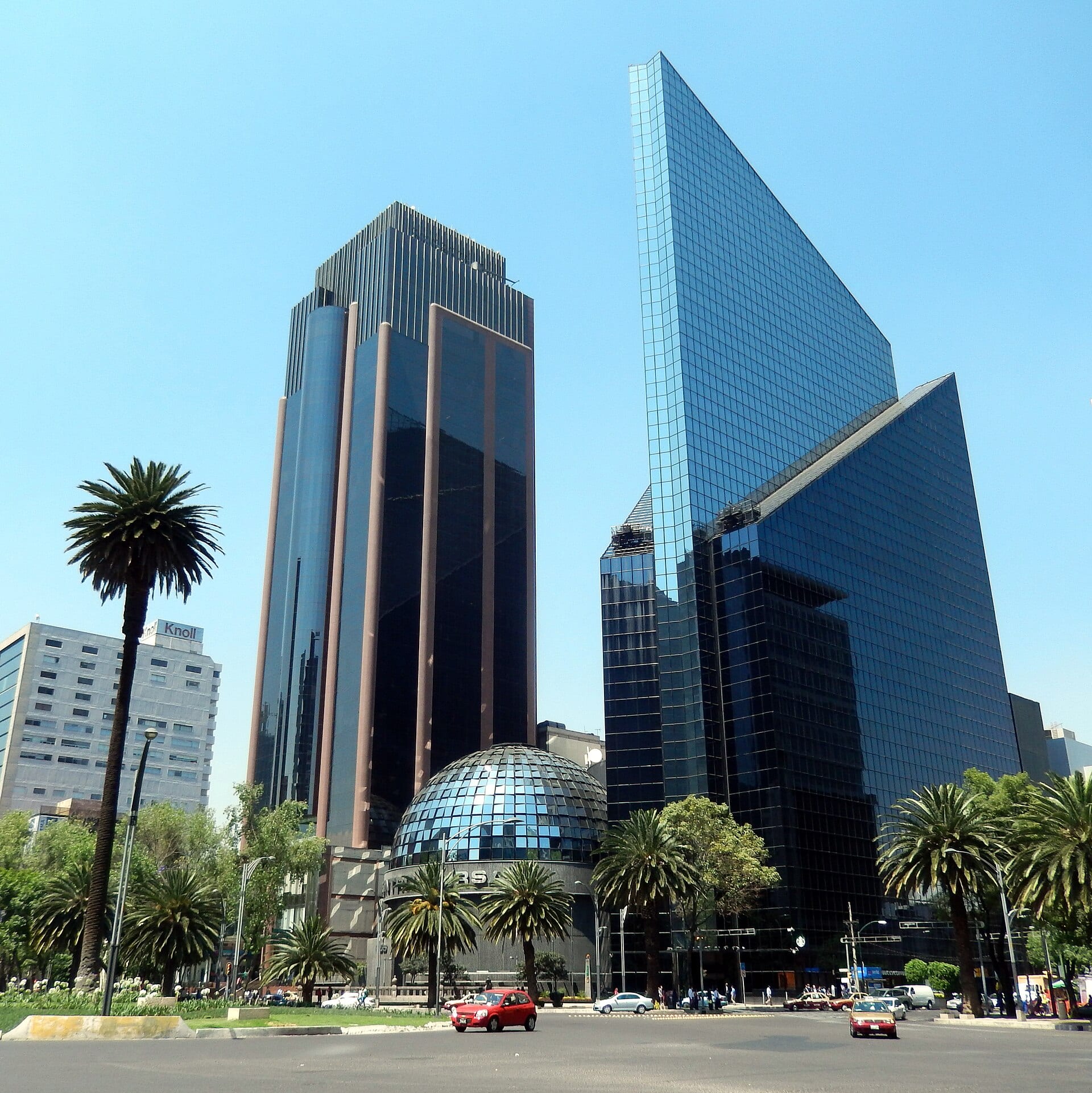 Dos rascacielos de cristal y la cúpula de la Bolsa Mexicana de Valores bajo un cielo despejado, con palmeras y autos en la avenida.