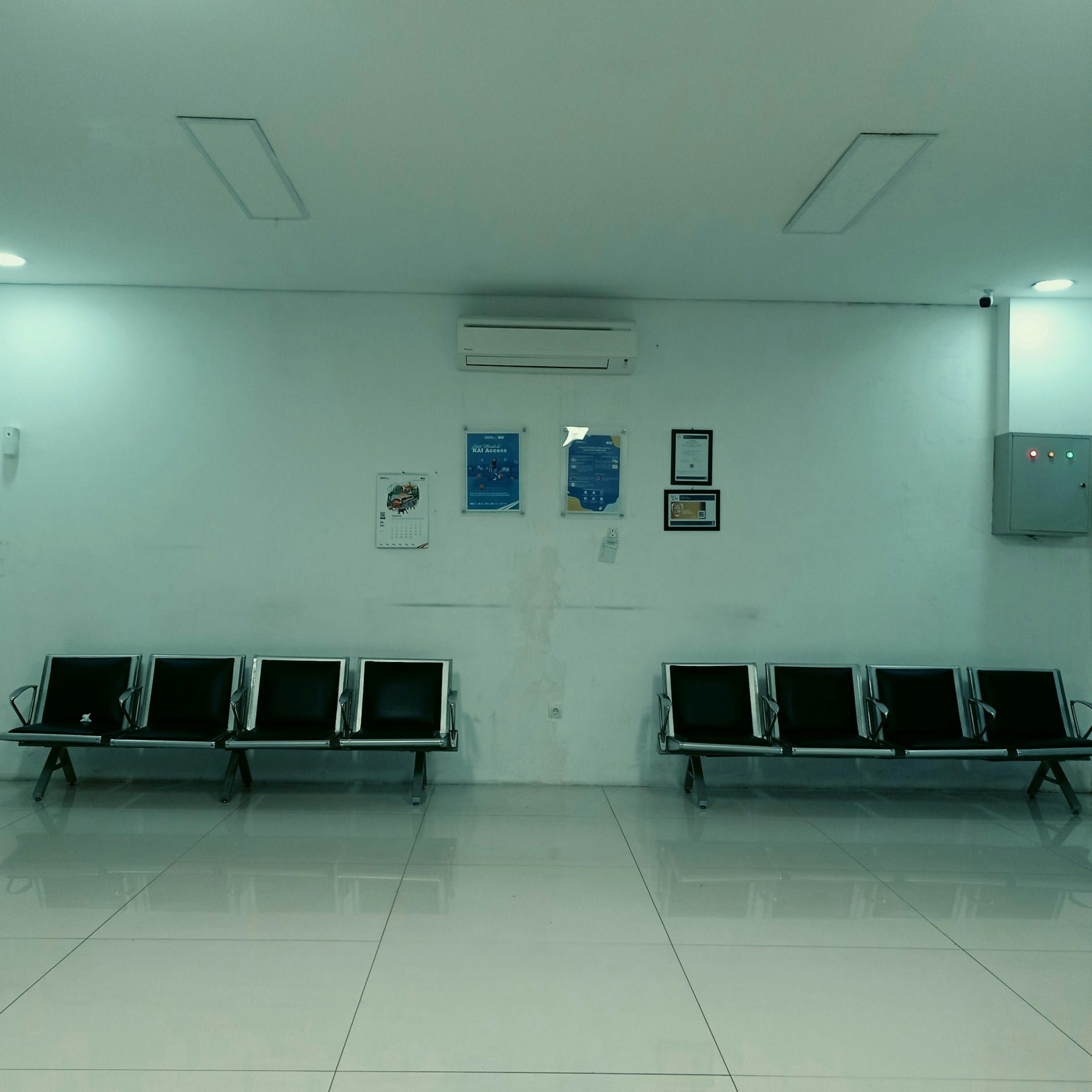 Dos hileras de sillas vacías de metal y asiento negro frente a una pared blanca, en una sala de espera con luz fría.