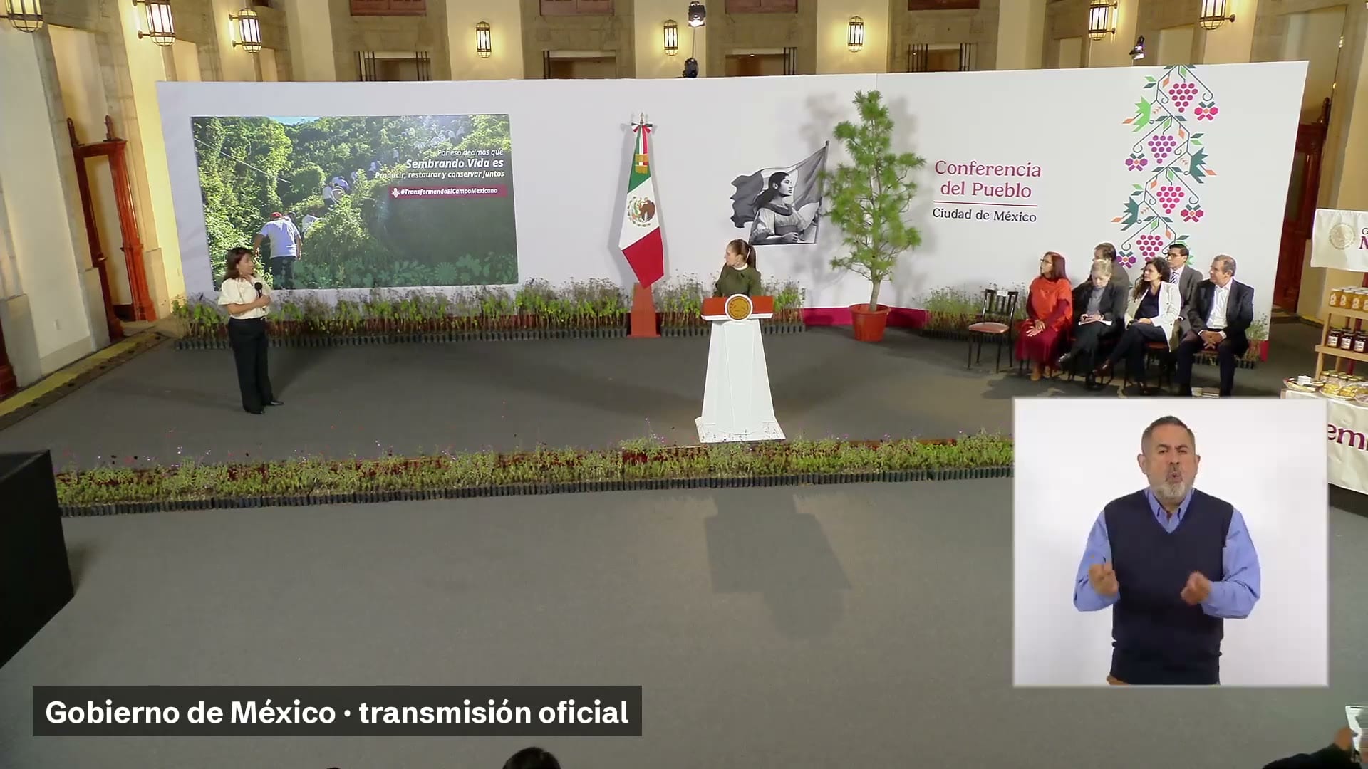 Una mujer habla junto a una pantalla con el lema de Sembrando Vida, mientras la presidenta escucha en el podio de la conferencia matutina.