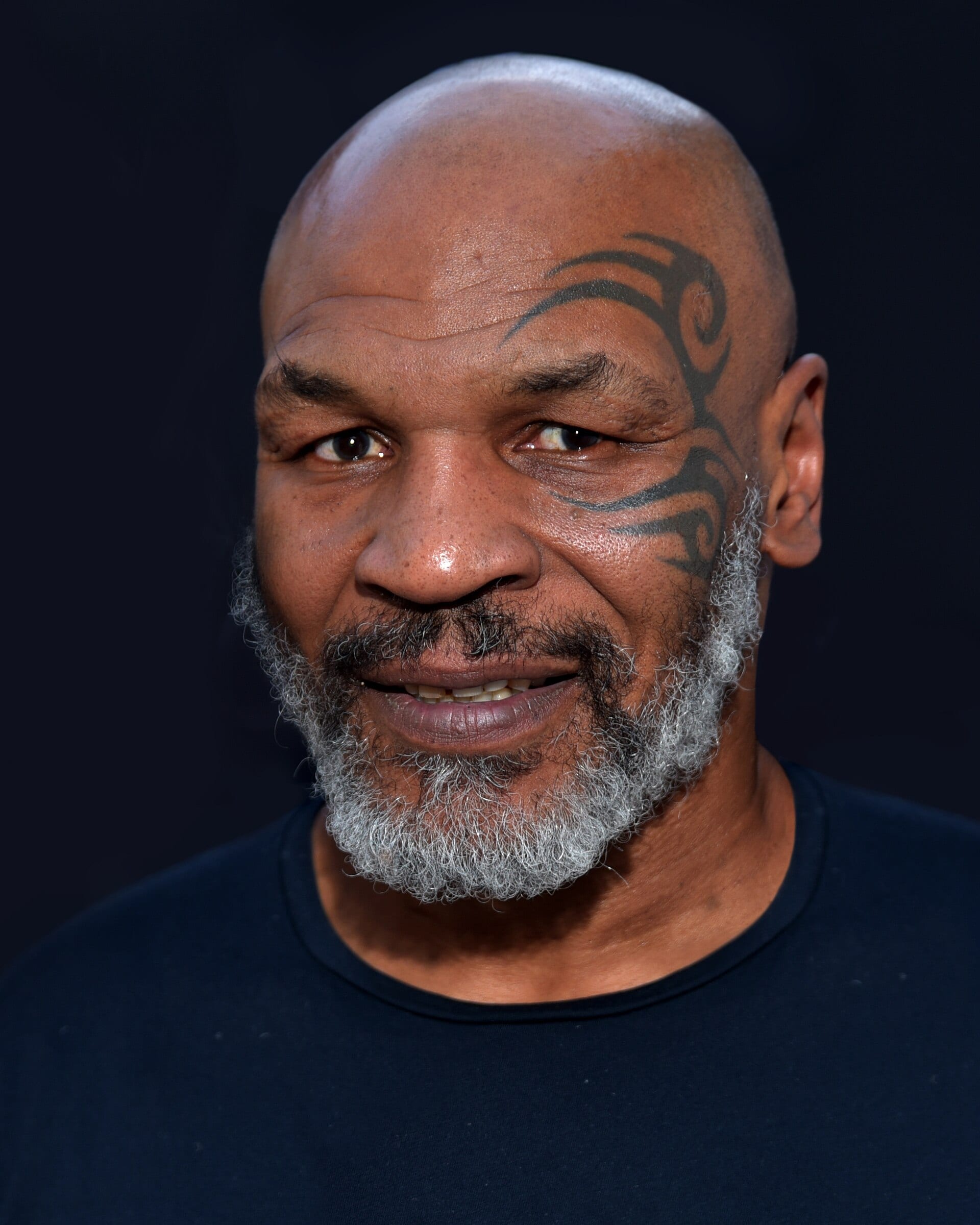 Retrato de Mike Tyson, con barba entrecana y el tatuaje tribal en el lado izquierdo del rostro, frente a un fondo oscuro.