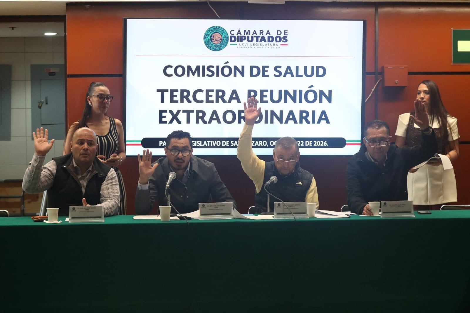 Cuatro integrantes de la Comisión de Salud levantan la mano para votar, sentados tras una mesa con mantel verde y bajo una pantalla que dice Tercera reunión extraordinaria.