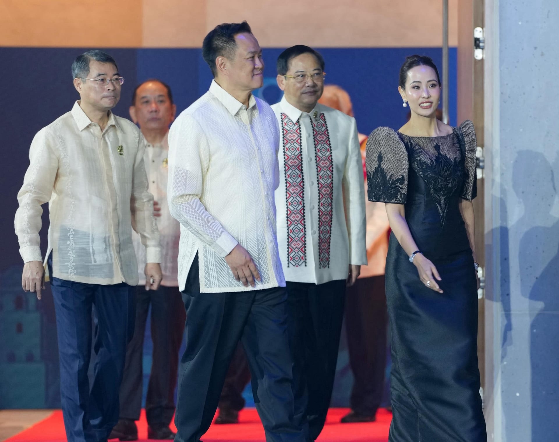 Four men in embroidered fiber shirts and a woman in a black dress with puffed sleeves walk along a red carpet inside a hall.