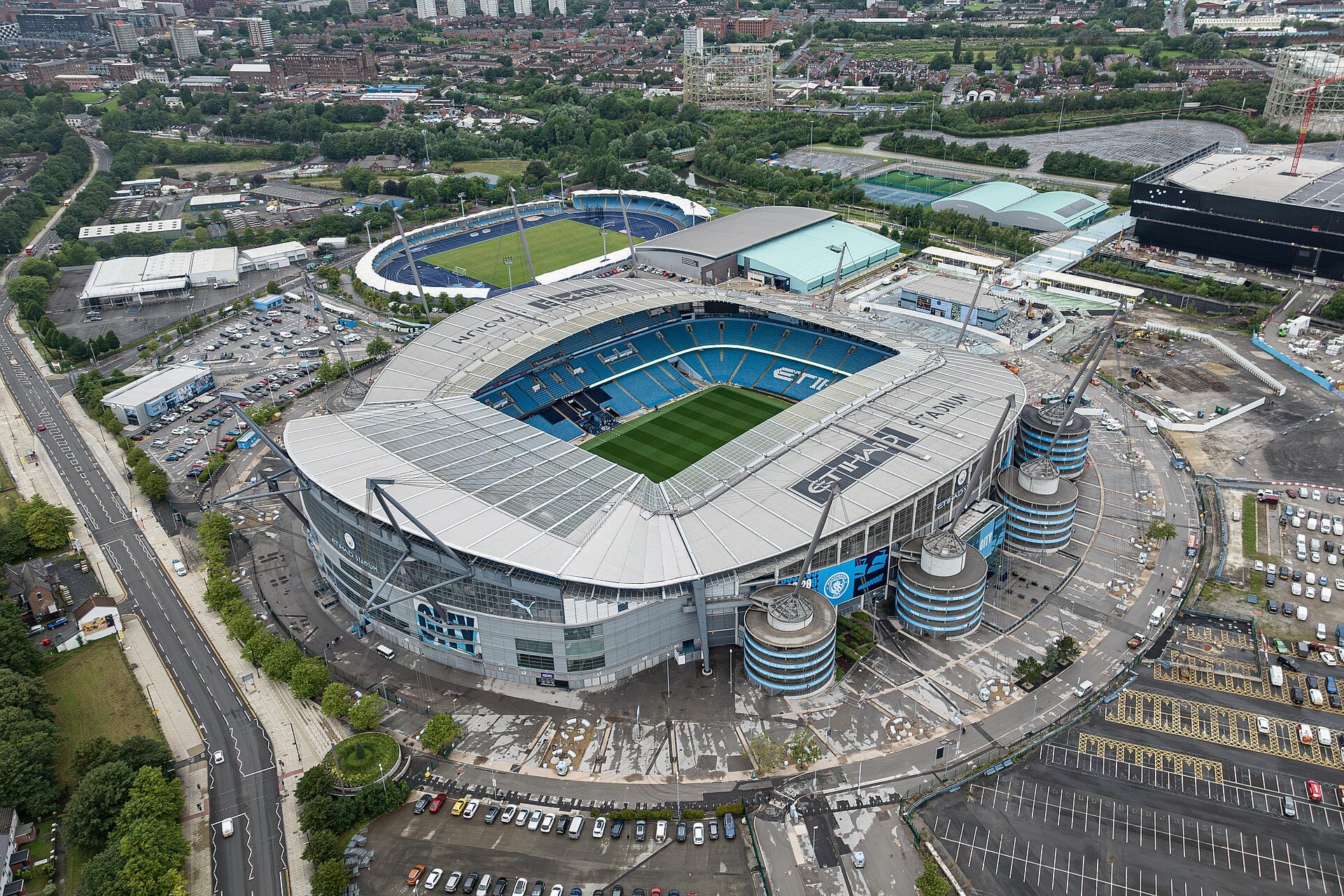 Vista aérea del Etihad Stadium de Manchester, con su techo gris, las gradas azules y los estacionamientos alrededor.