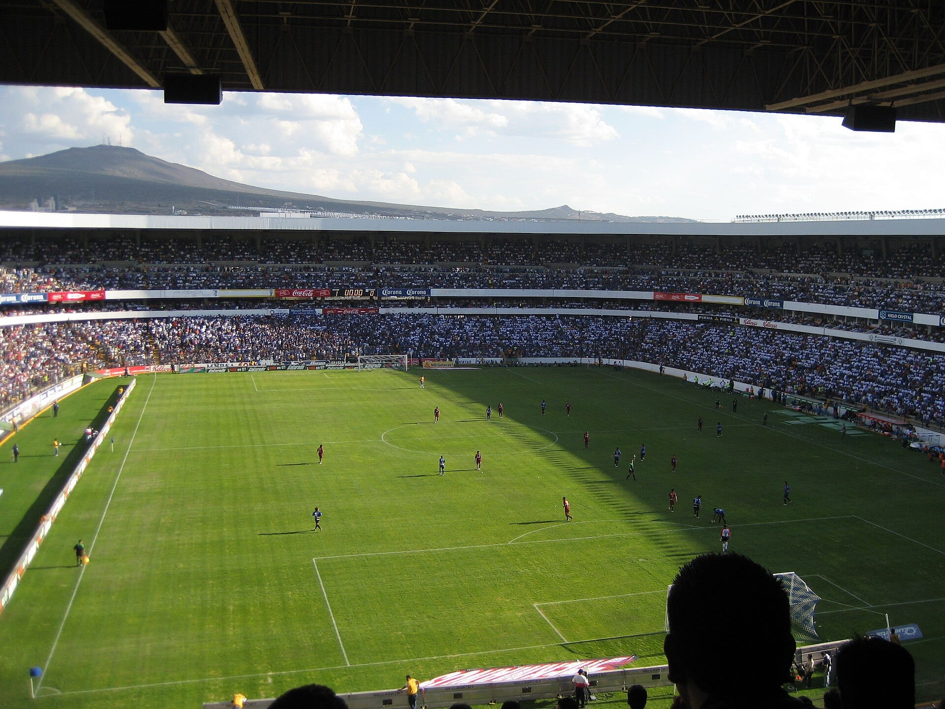 Vista desde la grada alta del Estadio La Corregidora con la cancha de pasto, jugadores dispersos y las tribunas llenas bajo un cerro al fondo.