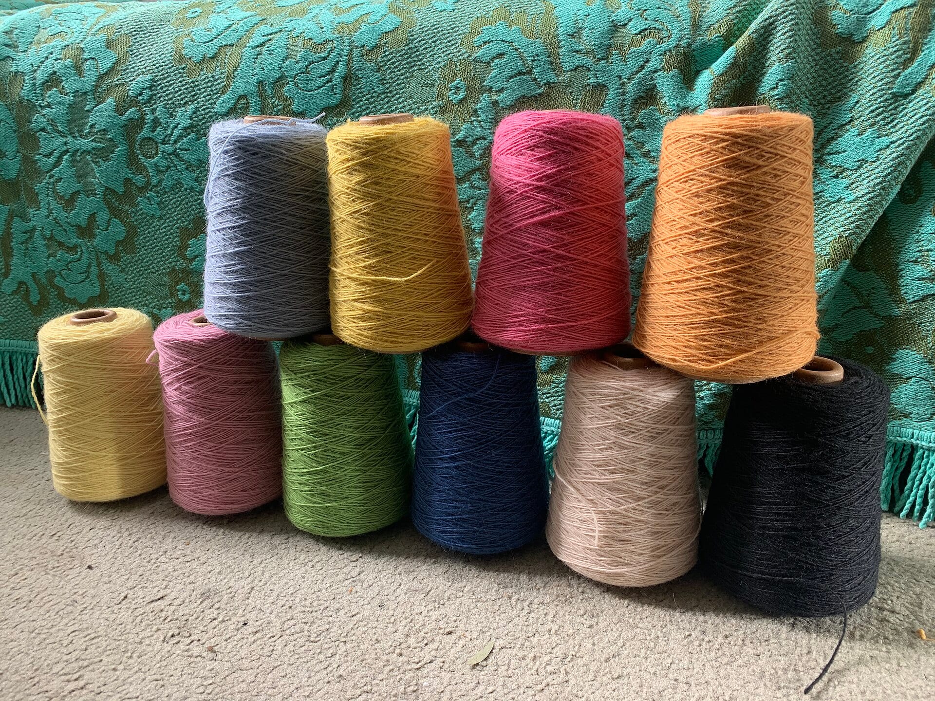 Diez conos de hilo de colores, apilados en dos filas frente a una tela verde con flores.