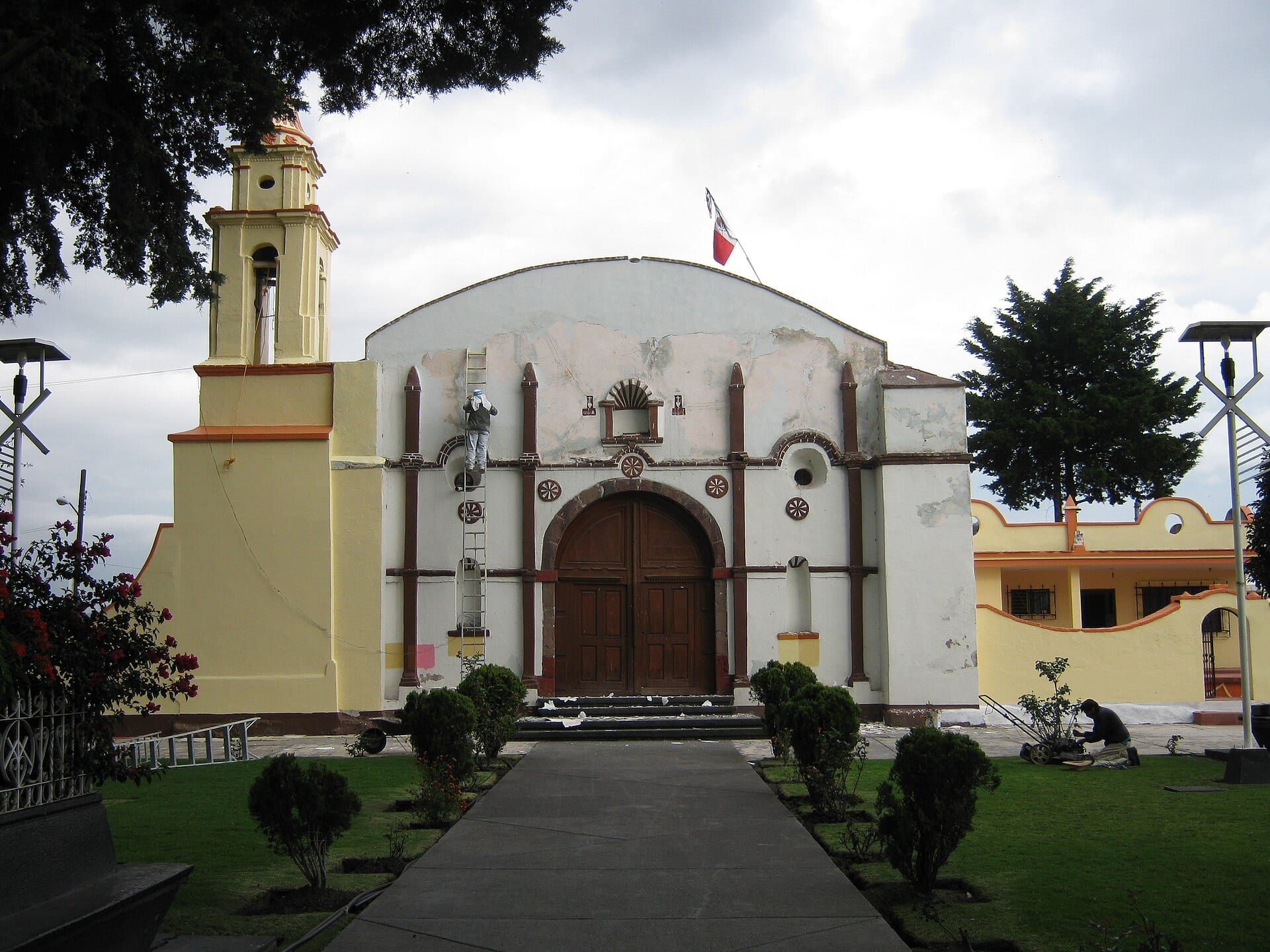 Fachada blanca de una iglesia con campanario amarillo y puerta de madera, frente a un jardín con andador.