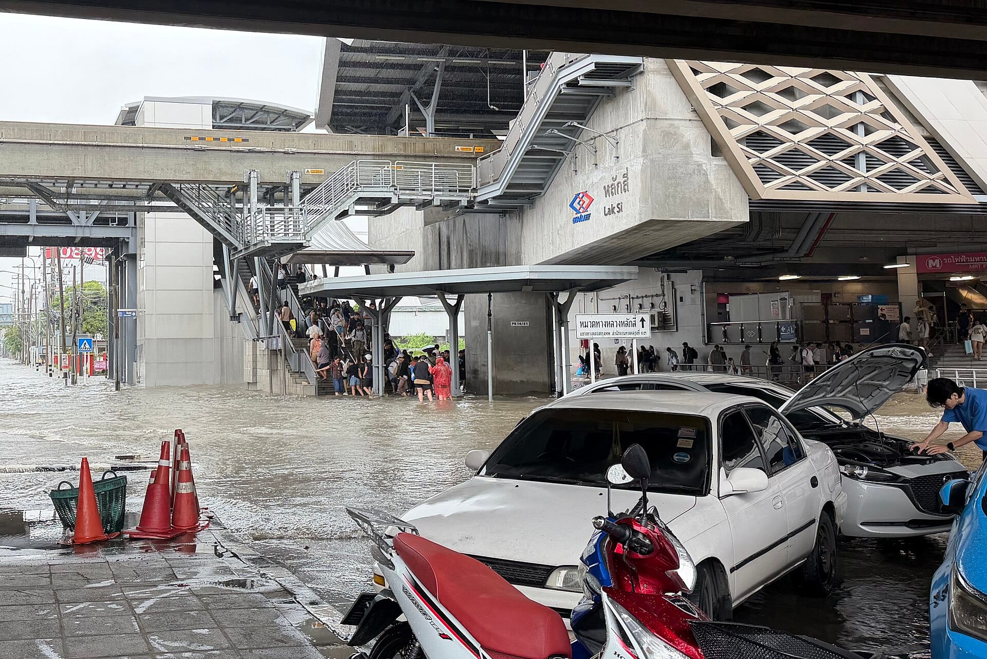 Una calle de Bangkok inundada junto a la estación Lak Si, con autos detenidos y gente bajando por una escalera.