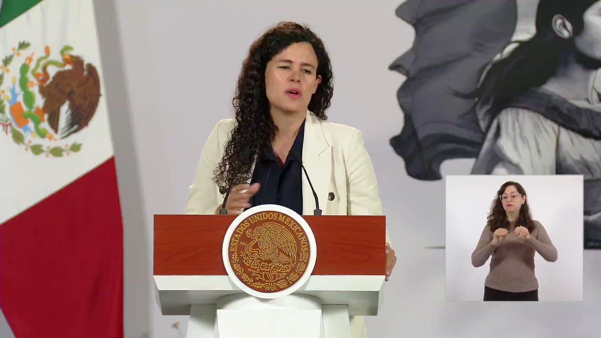 Una mujer de cabello rizado y saco blanco habla detrás de un podio con el escudo nacional, con una intérprete de lengua de señas en un recuadro.