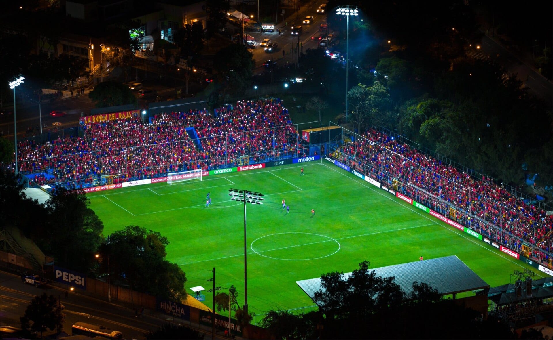 Vista aérea nocturna del estadio Manuel Felipe Carrera, con las tribunas llenas de aficionados con luces de celular y el campo iluminado.