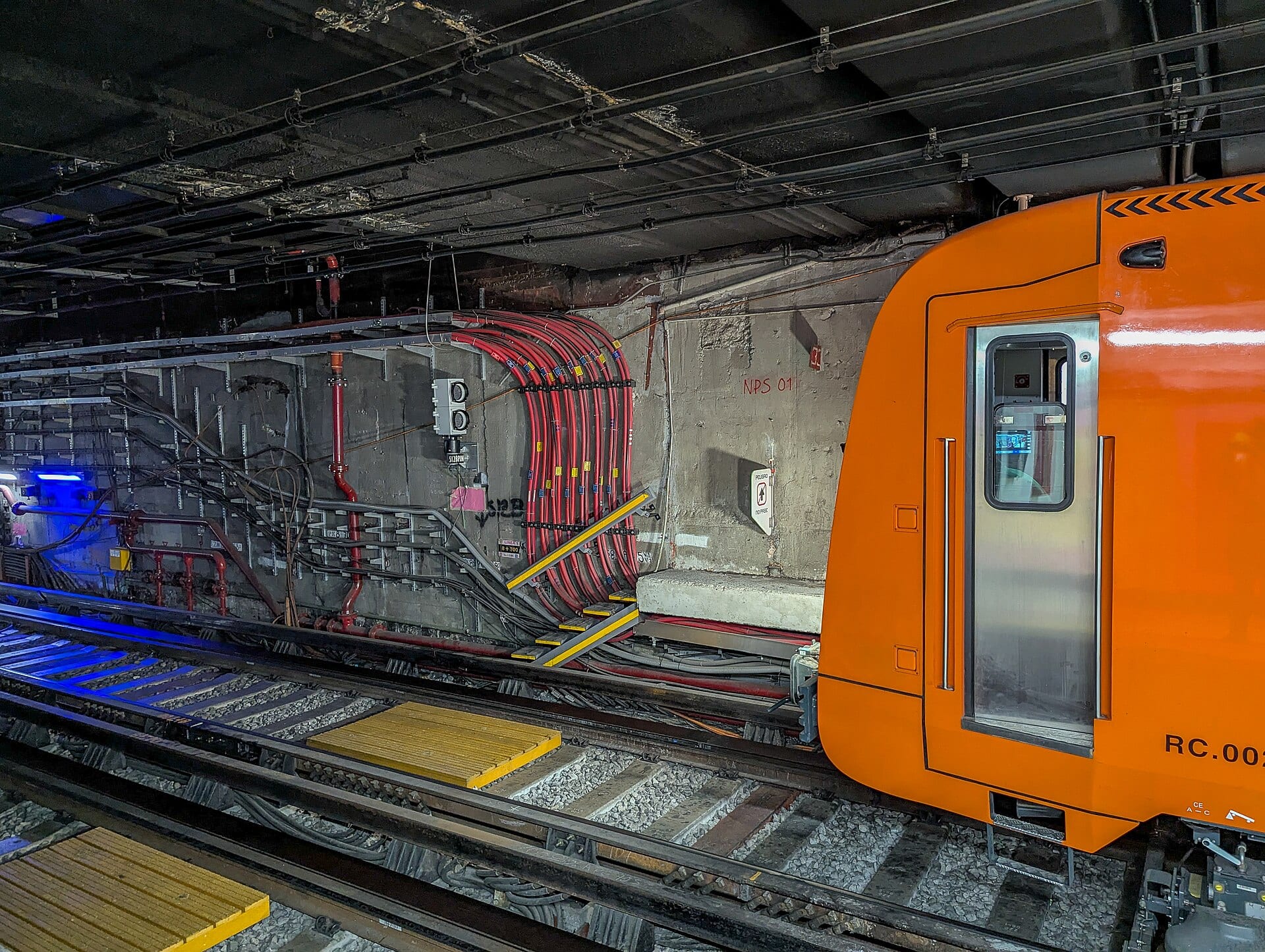 Un tren naranja del Metro con la puerta cerrada, detenido en un túnel junto a cables rojos y una pared de concreto.
