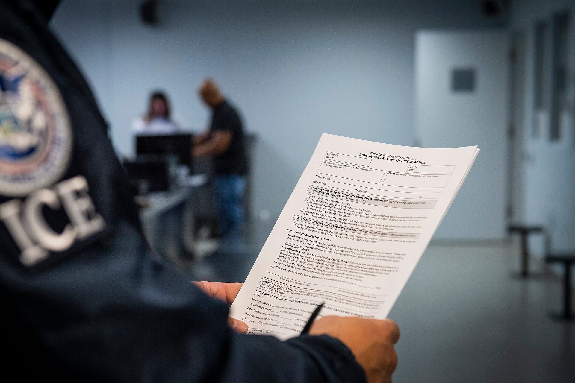 Un agente con el parche de ICE en el hombro sostiene un formulario de detención migratoria en una sala de un centro de detención.