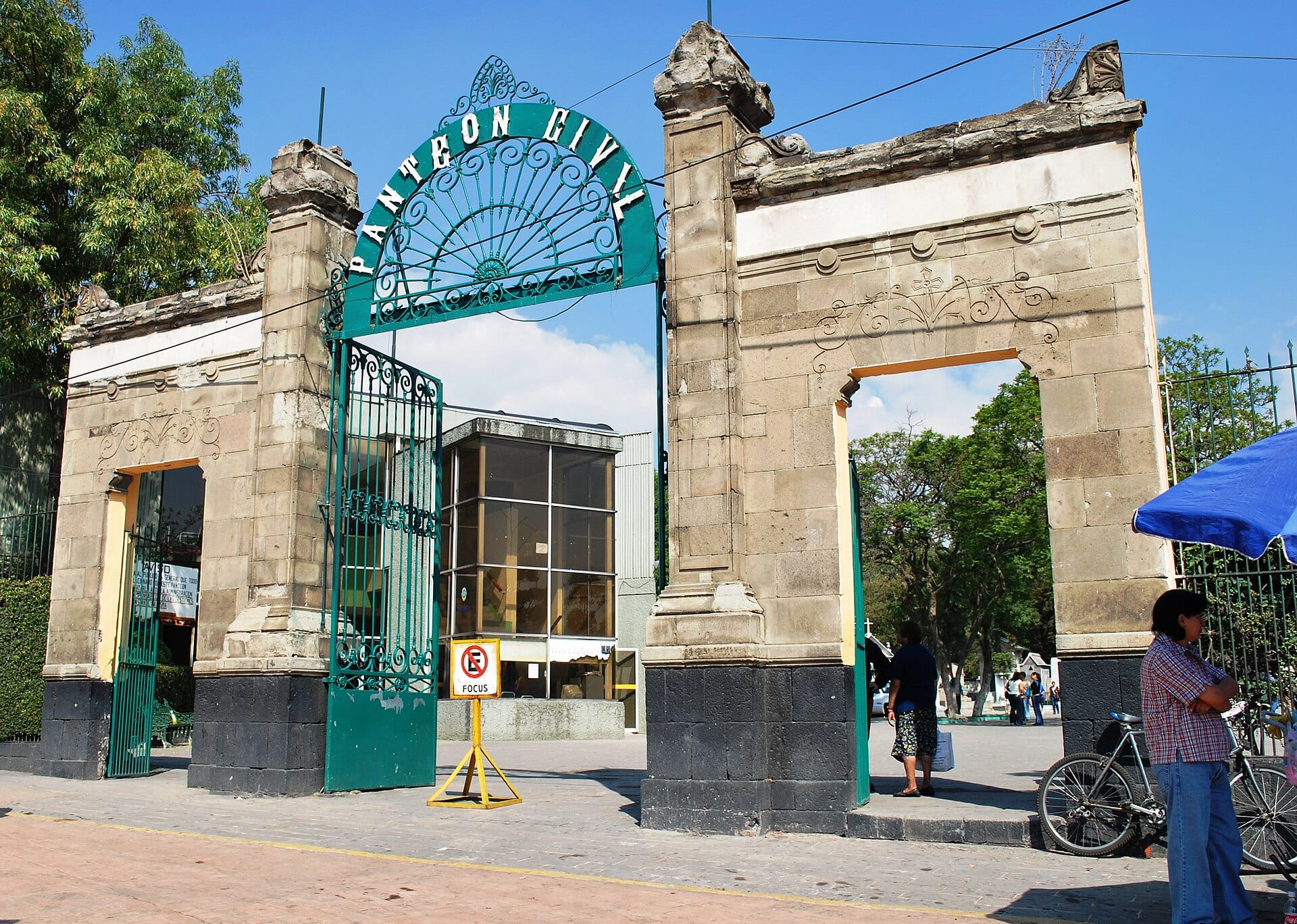 Entrada del Panteón Civil de Dolores, con un arco de herrería verde que lleva el nombre del cementerio entre dos pilares de piedra, y peatones junto a una bicicleta.