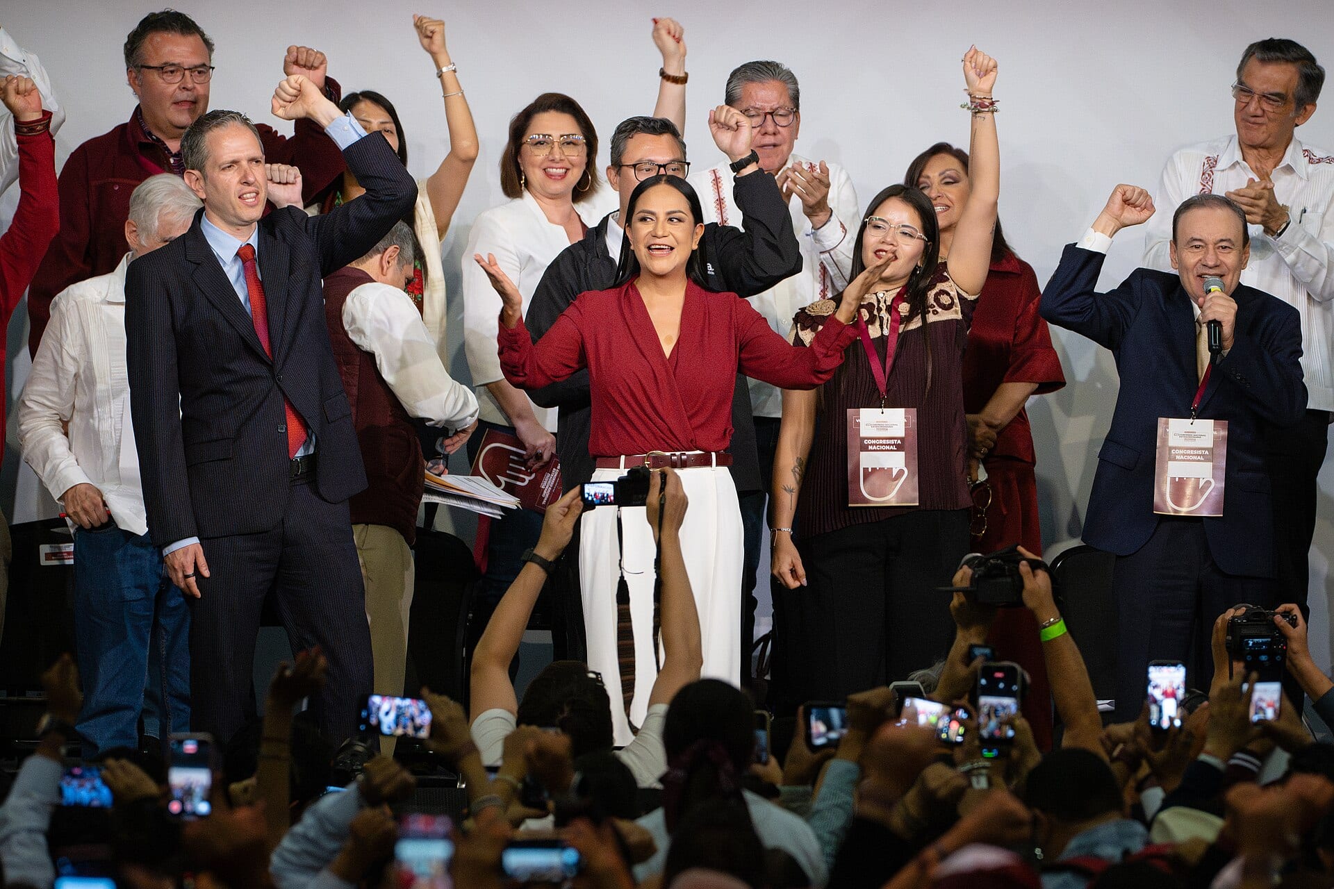 Una mujer de blusa roja extiende los brazos en un escenario, rodeada de personas con el puño en alto, ante un público que graba con celulares.