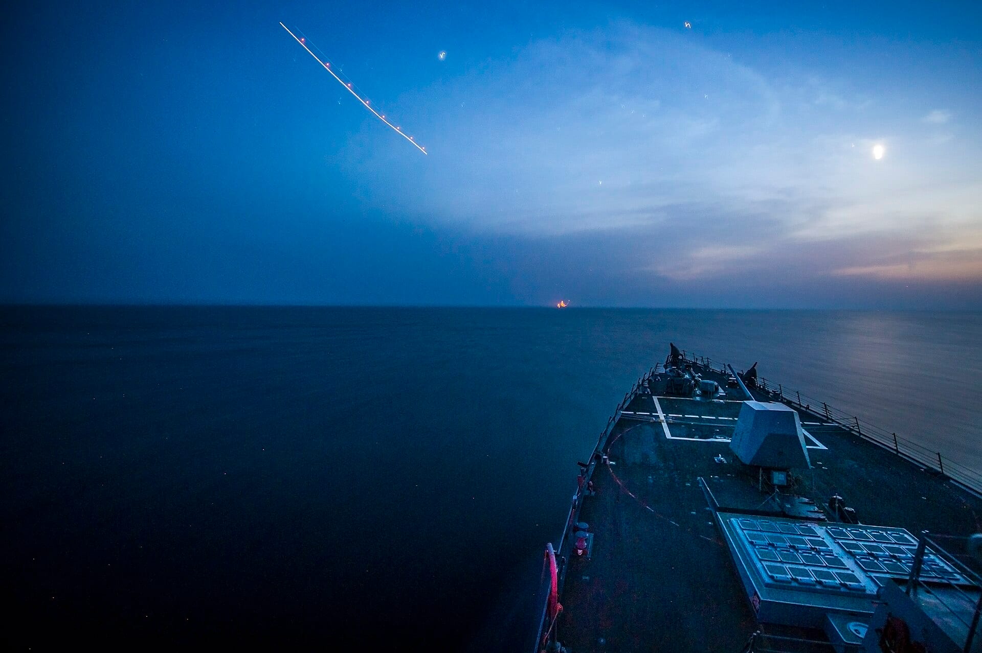 The bow of a US destroyer cruises at dusk on calm water, with the light trail of an aircraft in the sky.